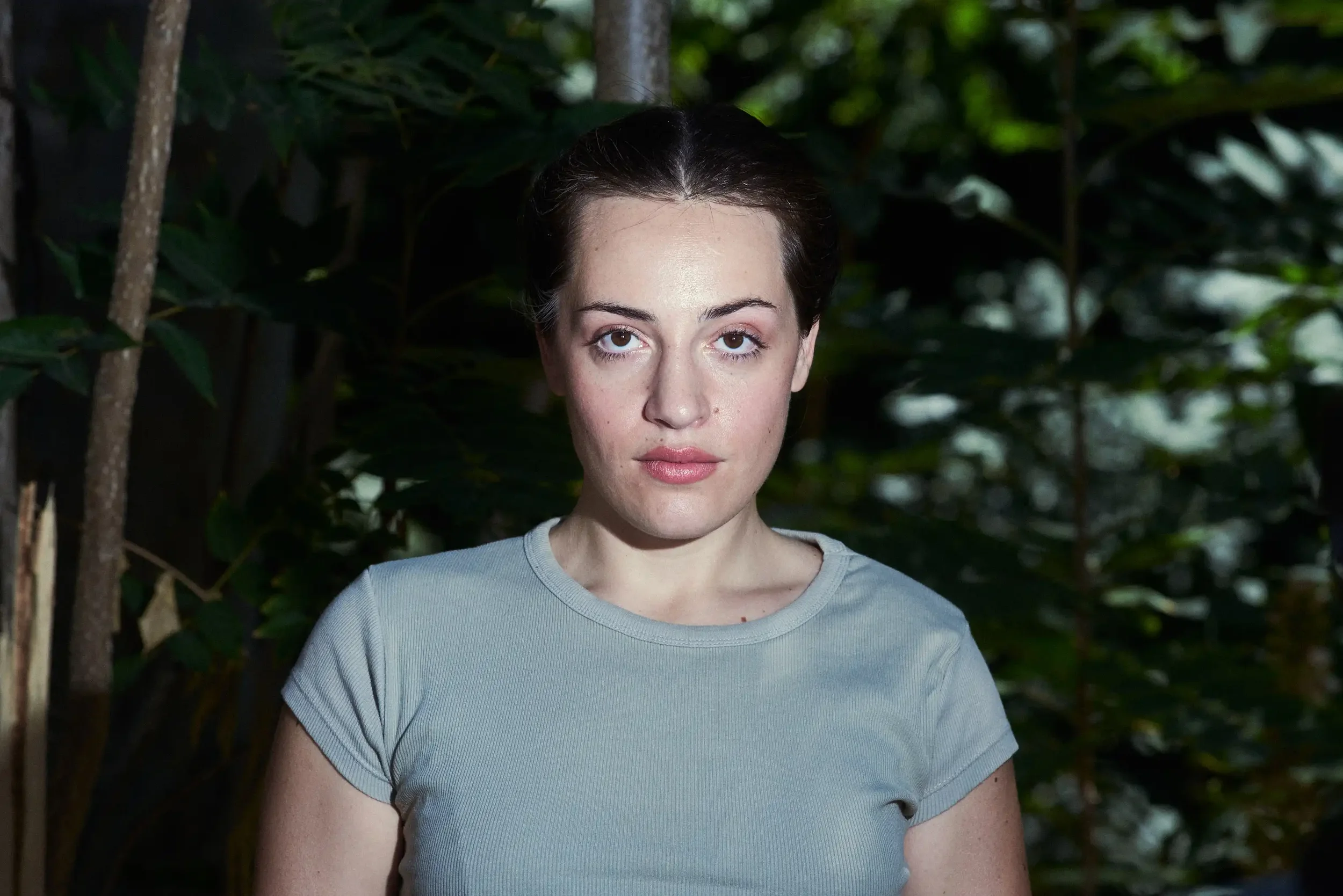 A woman with dark hair pulled back, wearing a light gray t-shirt, standing outdoors in front of a background of dense green foliage.