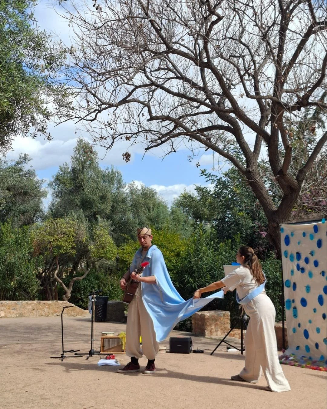 A man playing guitar and a woman holding a flowing piece of fabric outdoors, with trees and a blue patterned backdrop in the background.