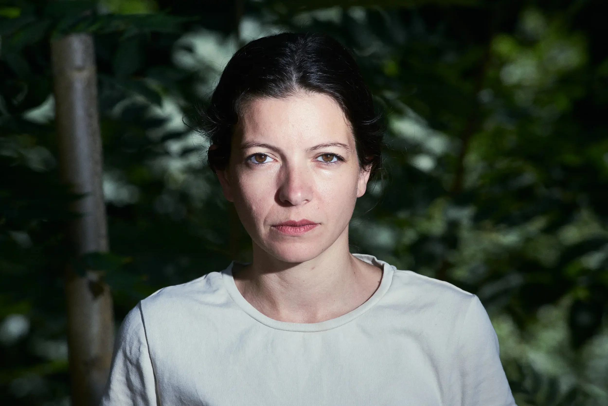 A woman with short dark hair standing outdoors in front of green foliage, wearing a white shirt.