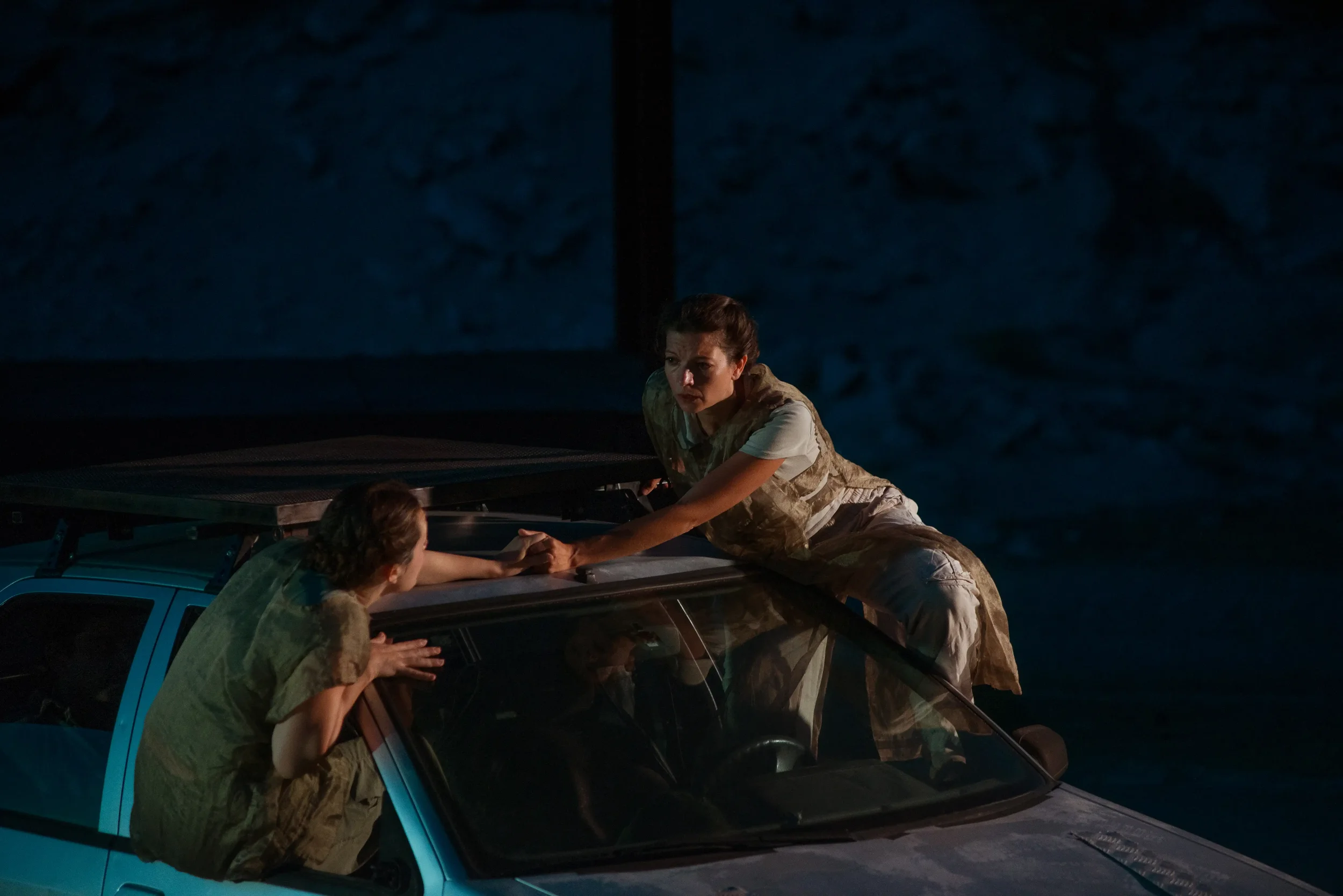 Two women in military camouflage outfits are on top of a car, one reaching out and holding the other's hand. It appears to be nighttime or in a dark setting.
