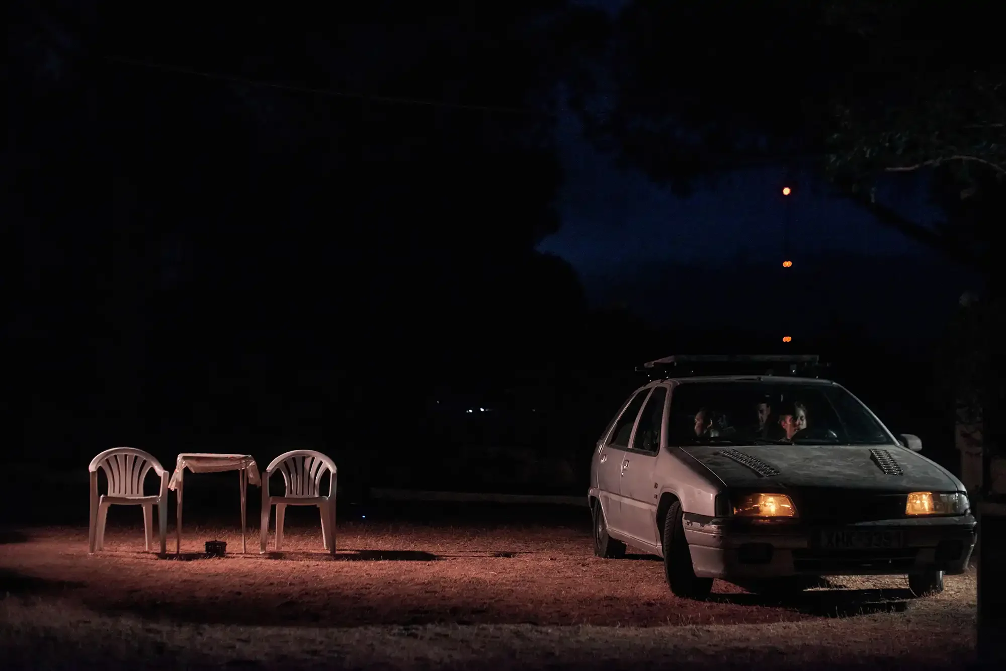 A car parked outside at night with two plastic chairs and a small table nearby, illuminated by a soft light.