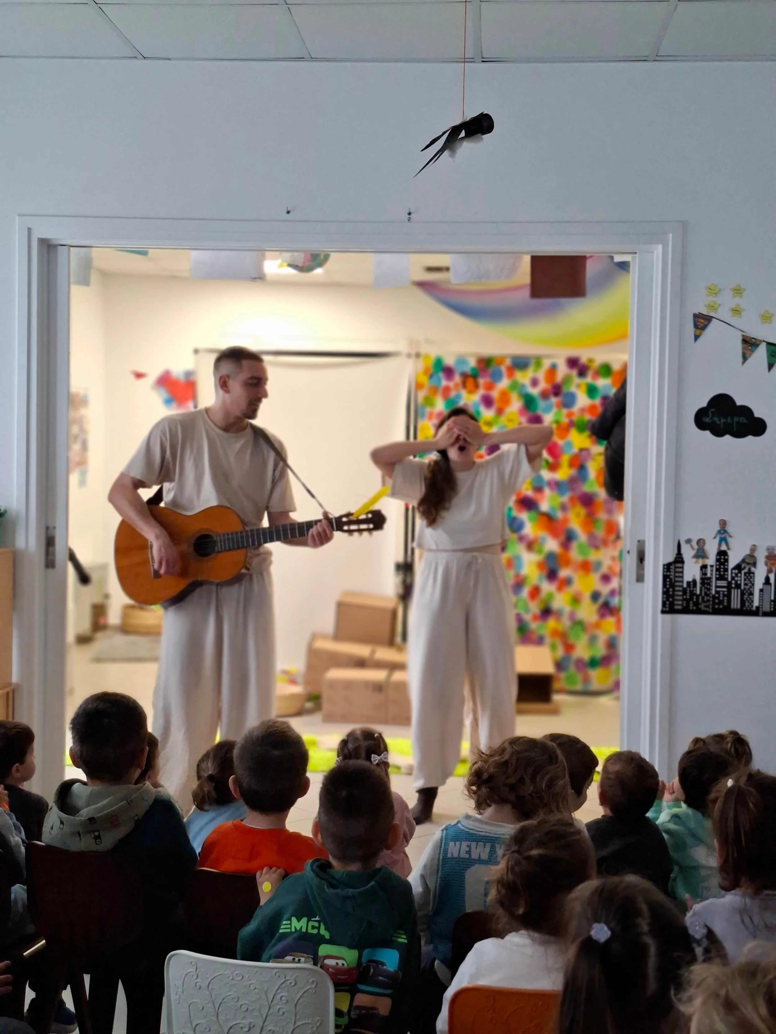 Children sitting on the floor watching a performance in a classroom with colorful walls, two performers on stage playing guitar and singing, dressed in white, with a rainbow decoration behind them.