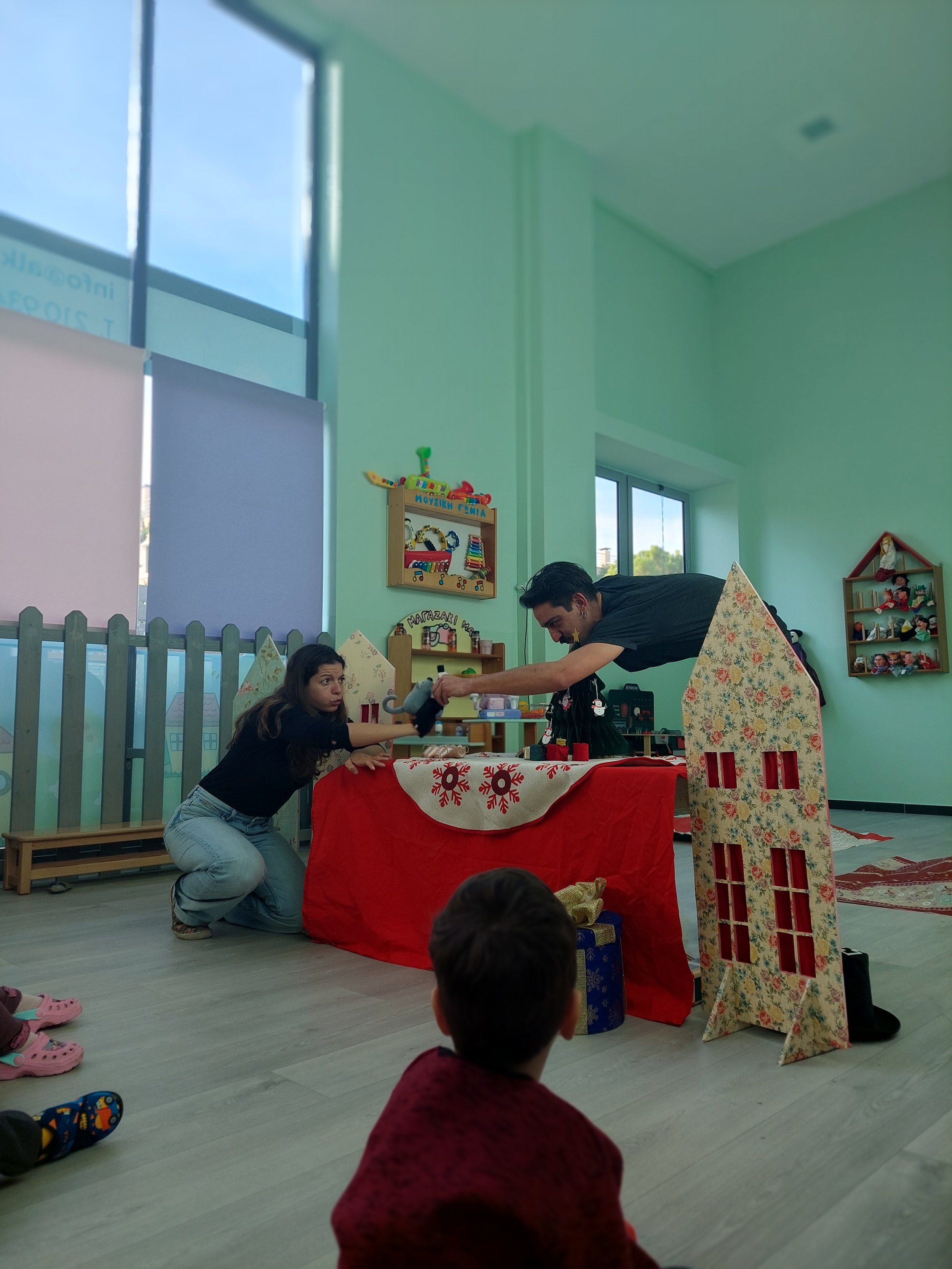 A man and a woman performing a puppet show or play for children in a room decorated for Christmas, with children watching.