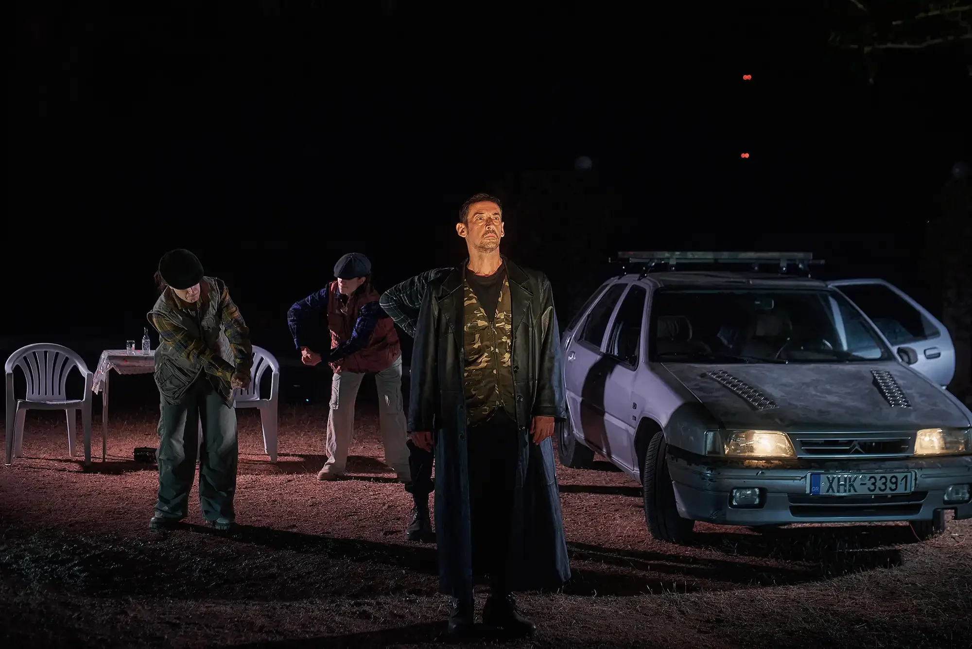 A scene at night with four people and two cars. One man stands in the foreground with a serious expression, wearing a leather jacket and camouflage shirt. Two women are adjusting something near a picnic table on the left. Two cars are parked behind t
