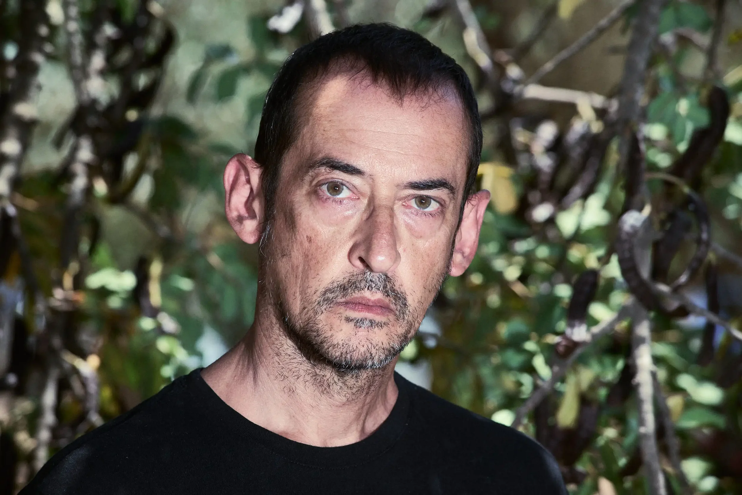 A man with short dark hair, light skin, and a beard, wearing a black shirt, looking serious with a background of leafy branches.