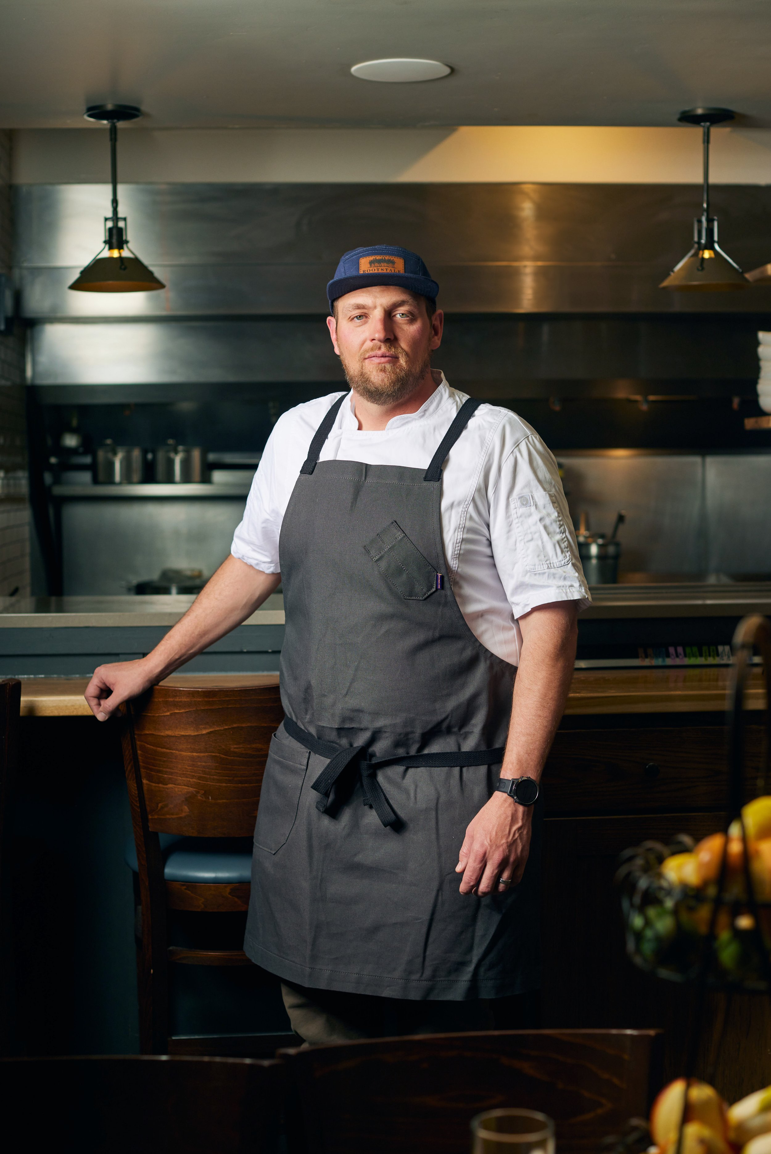 A smiling man with a beard and blue eyes, wearing a blue plaid shirt and a navy apron, standing indoors next to a white planter with a small tree. Books are visible on the left side of the image.