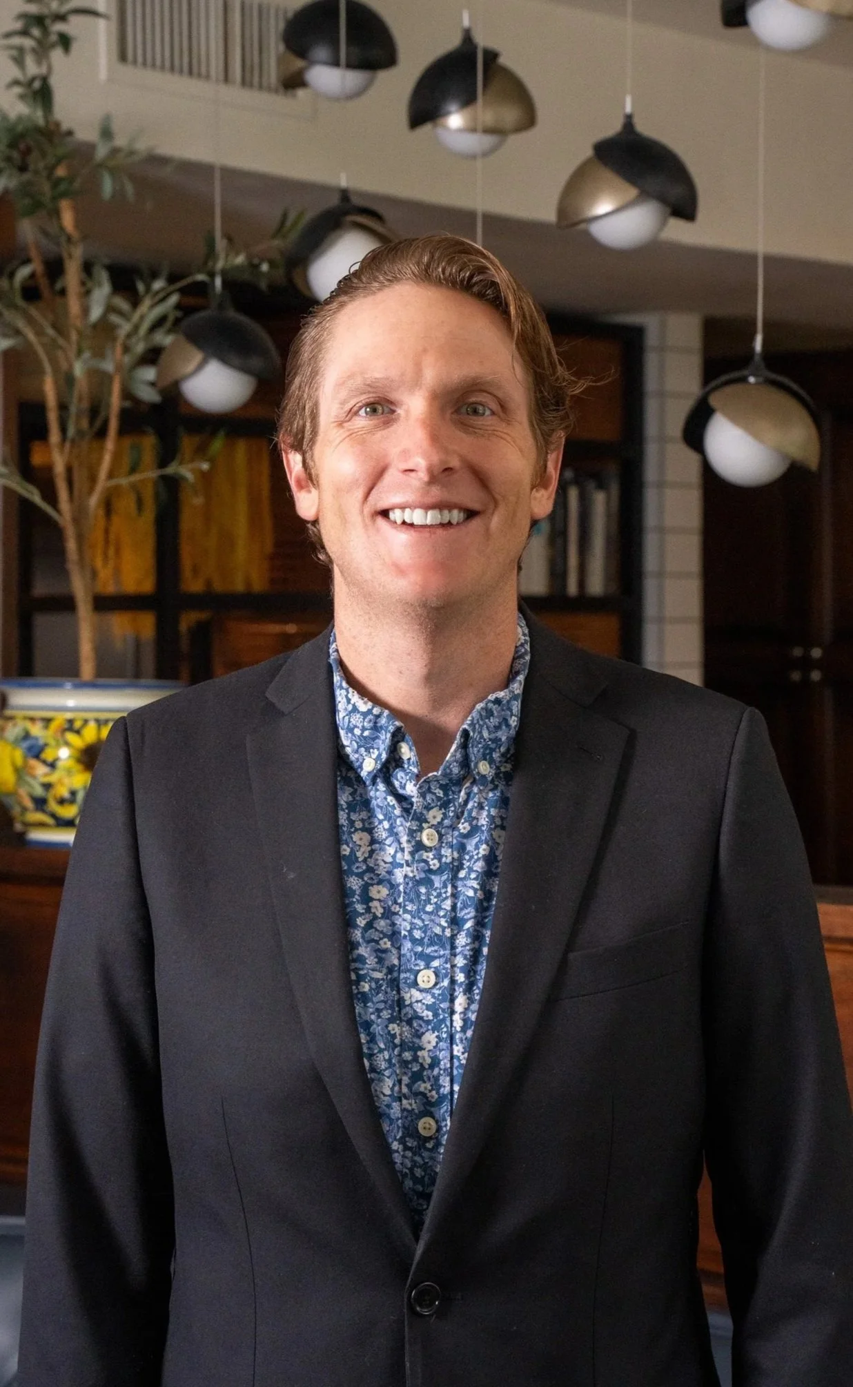 A young man with red hair, blue eyes, and a big smile, wearing a blue striped button-up shirt, standing in a bar with shelves of bottles in the background.