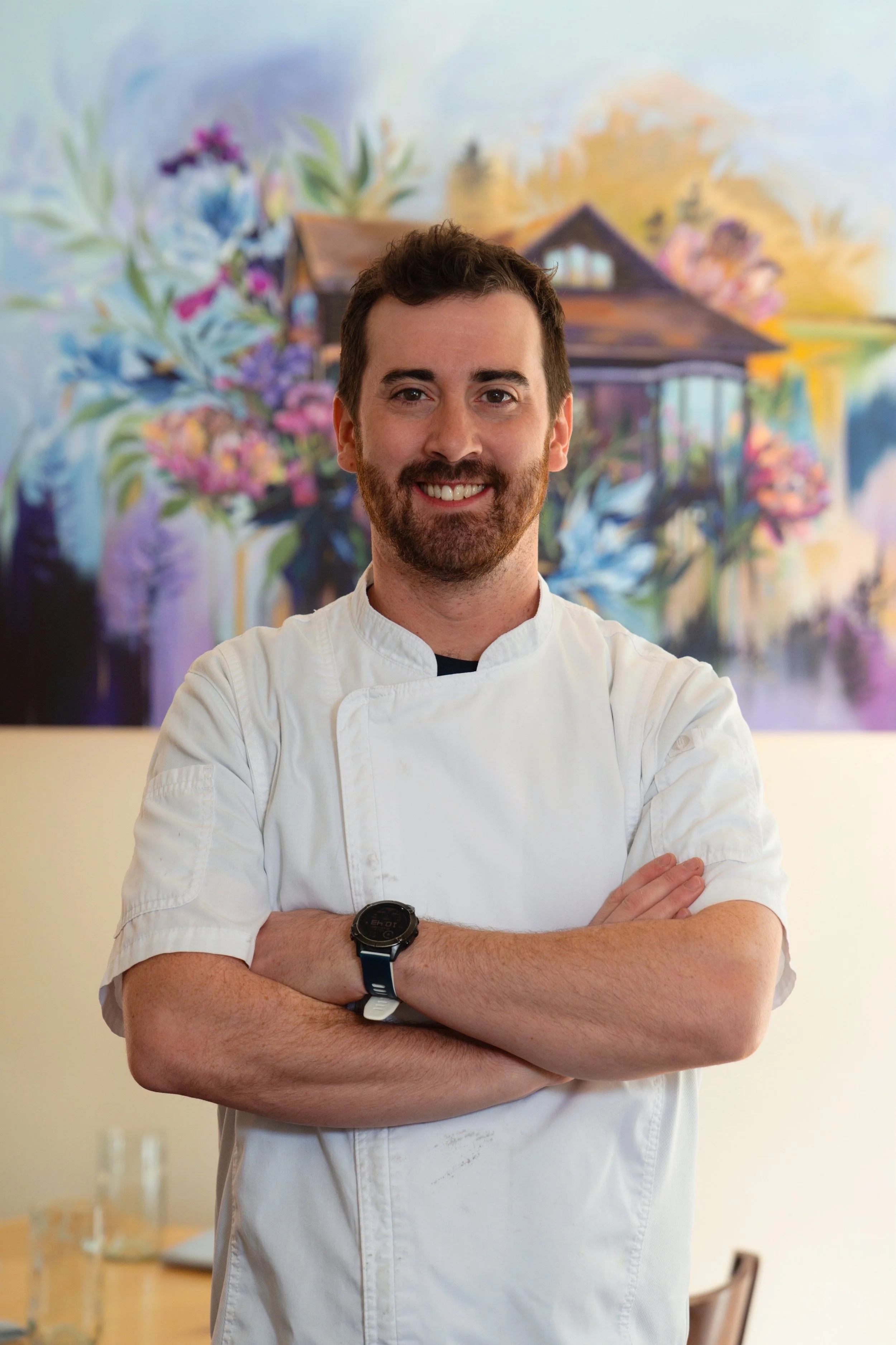 A man with a beard, wearing a black baseball cap backwards, a blue shirt, and a dark blue apron with pens in the pocket, smiling inside a flower shop or similar setting.