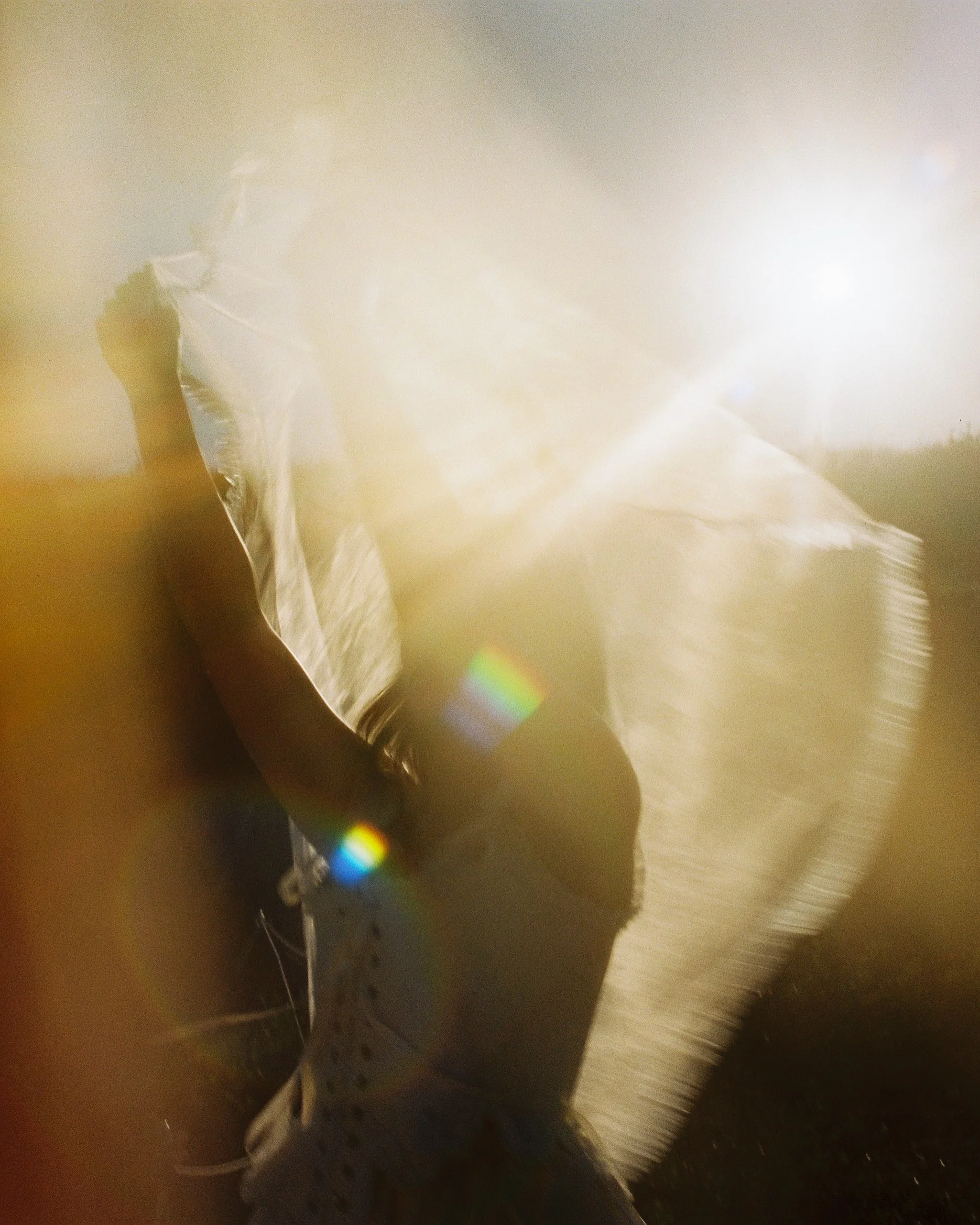 A person holding a large, partially transparent white plastic bag or sheet, with sunlight reflecting off it, creating a rainbow-like flare. The person's face is not visible, and they are dressed in patterned clothing with a dark baseball cap.