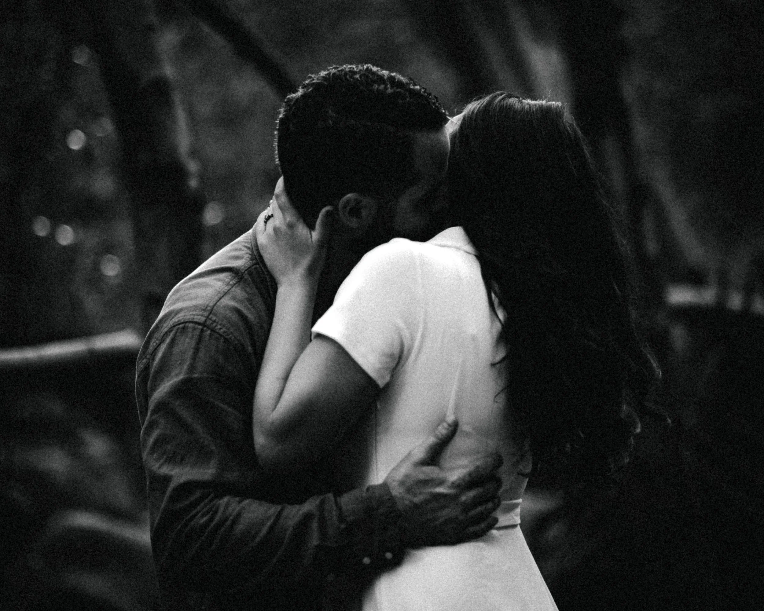 Moody, cinematic black and white photo of a couple kissing, with the man holding the woman's head and her hand on his neck, in an intimate embrace.