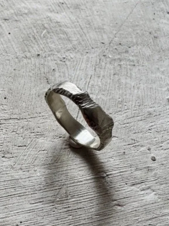 Handmade sterling silver band ring with organic edges, sculptural shape and raw textured surface on a concrete background.