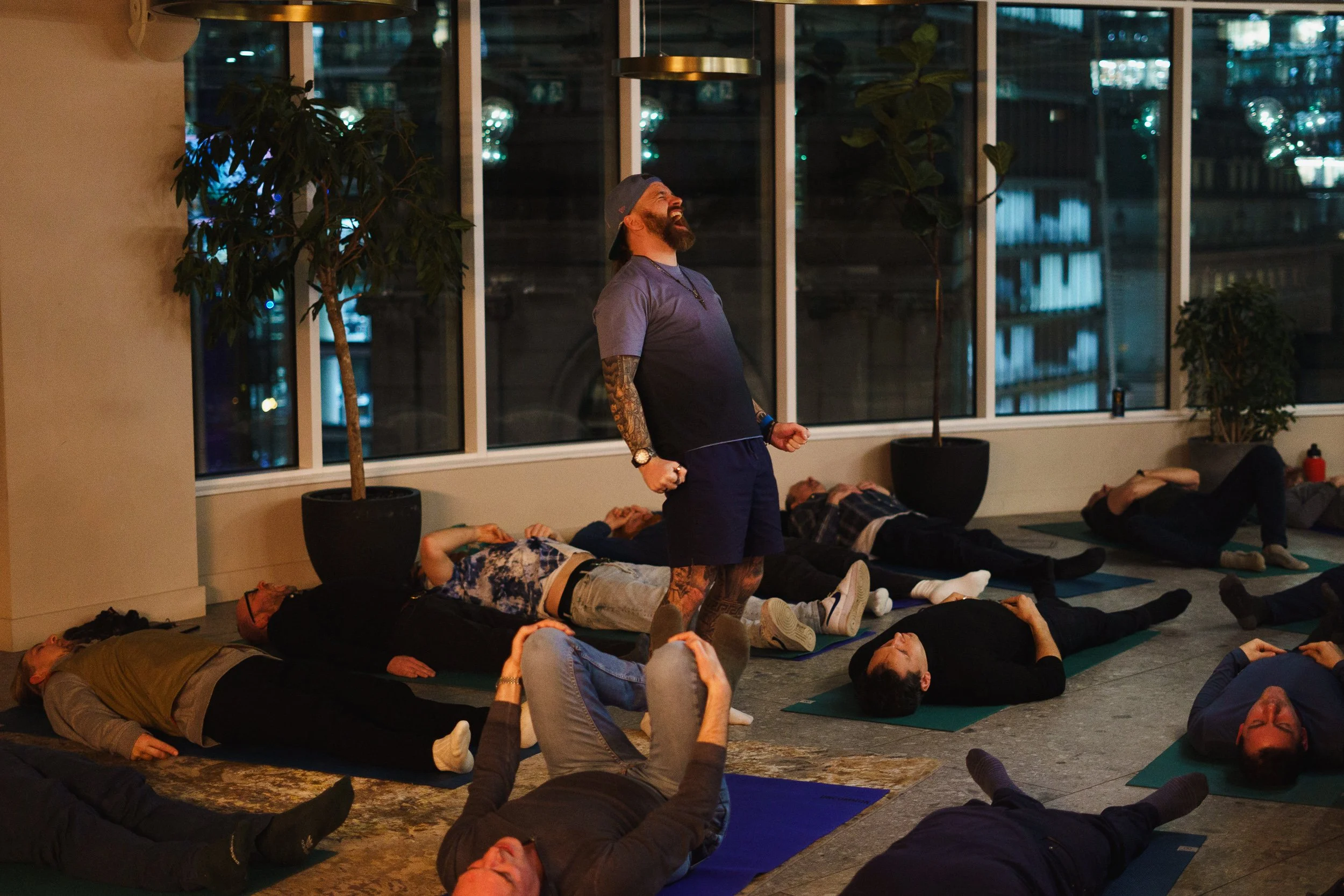 A group of people lying on yoga mats on the floor during a breathwork session at night in a room with large windows and indoor plants, with an instructor standing and laughing at the front.