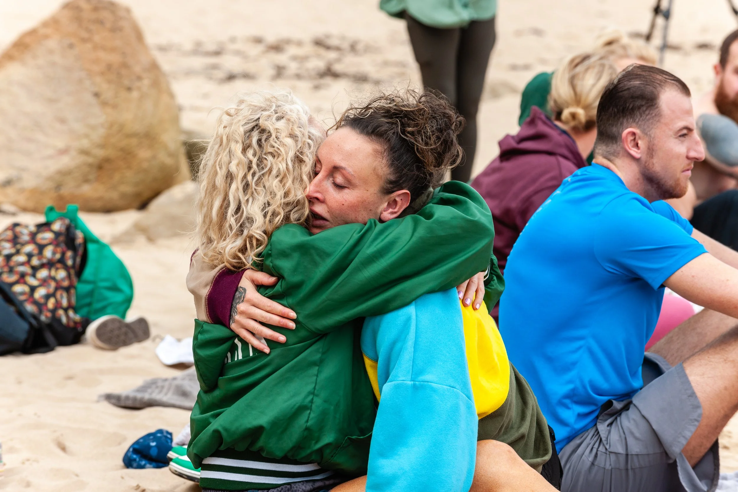 Two women hugging each other tightly on a beach while people sit nearby, some with their eyes closed and hands on their laps after a josh Connolly breathwork event on the beach