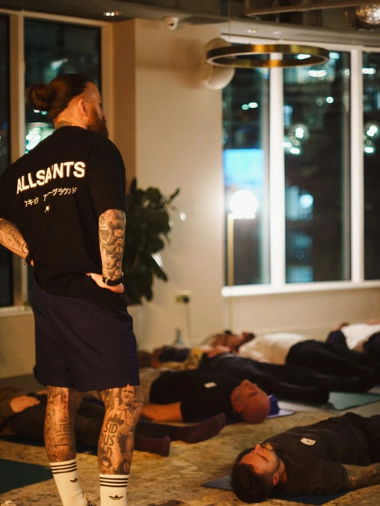 Josh Connolly standing in breathwork session, looking over people with their eyes closed, lying on mats on the floor, inside a room with large windows at night.