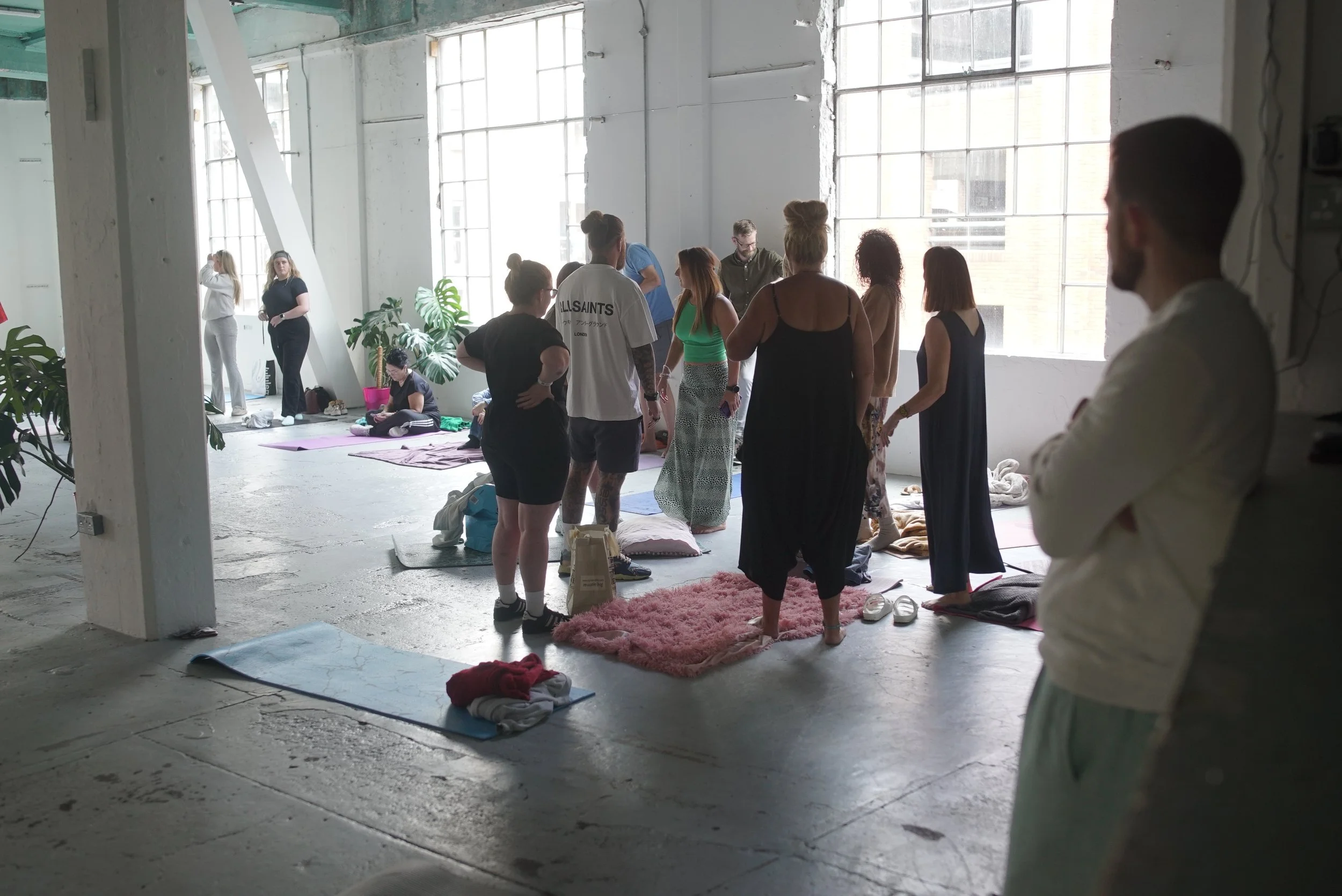 A group of people gather in a bright, industrial-style loft with large windows with Josh Connolly. Some are standing and chatting, while others sit on yoga mats or pillows on the floor. Various plants and personal belongings are scattered around.