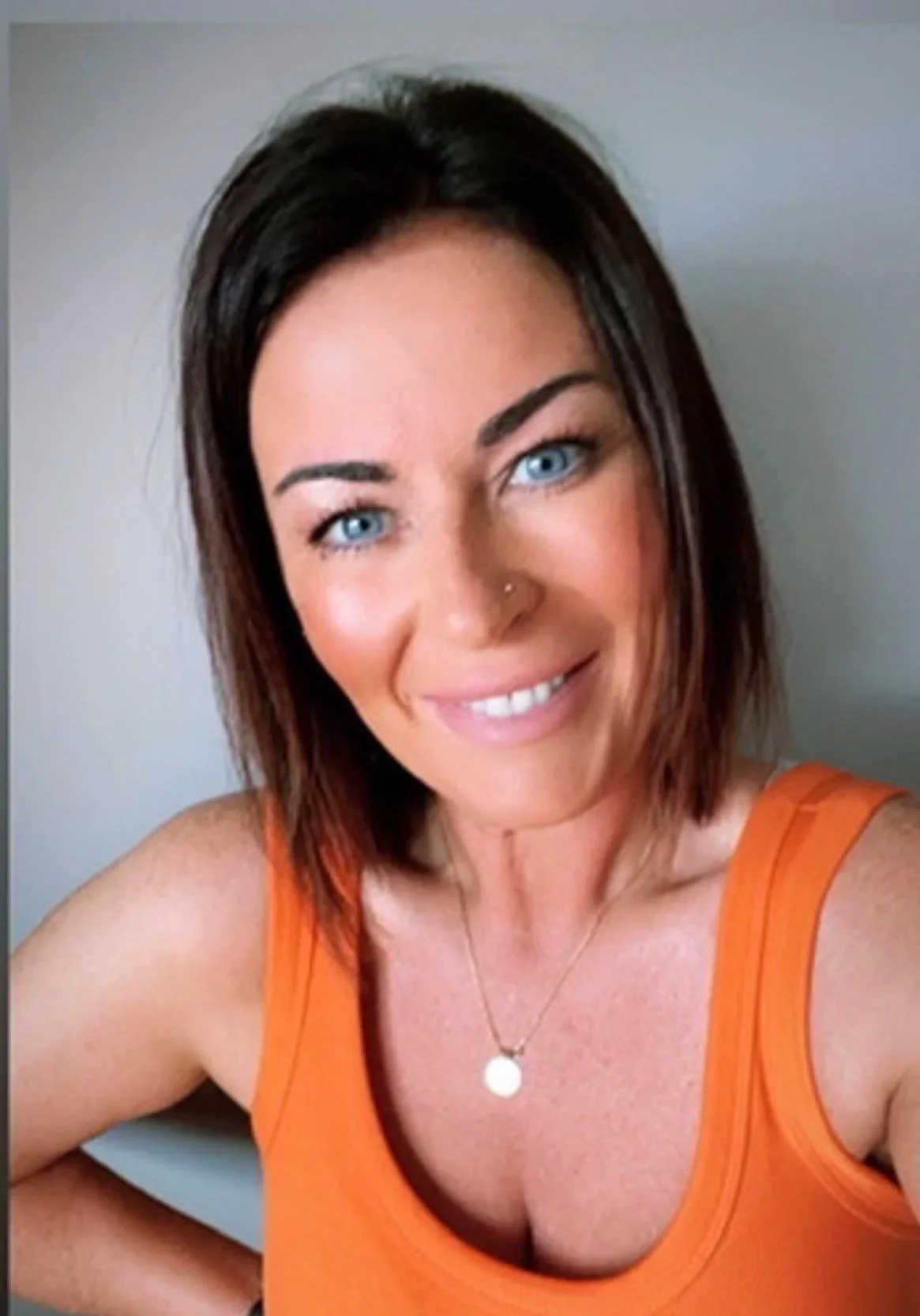 A woman with shoulder-length dark hair, blue eyes, and a nose piercing, smiling at the camera. She is wearing an orange sleeveless top and a gold necklace with a round pendant, against a light-colored background.