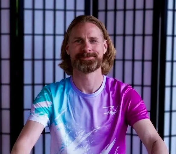 A man with long hair and a beard sitting in front of a black folding screen, wearing a colorful tie-dye shirt with shades of purple, pink, blue, and white.