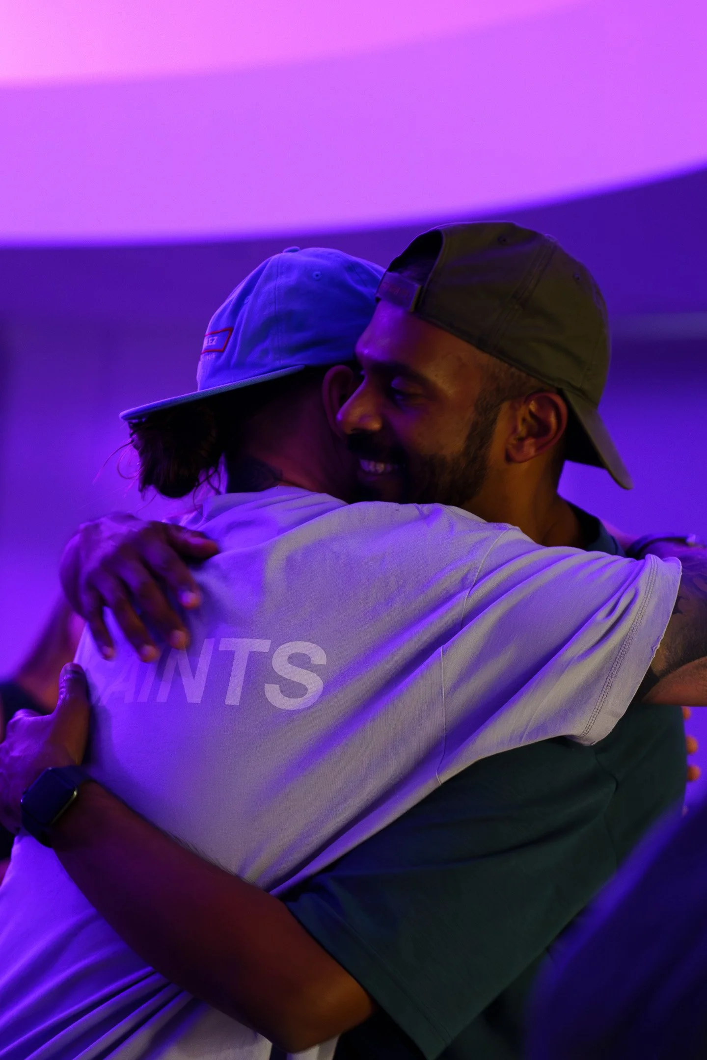 Josh Connolly hugging a friend, one wearing a white shirt, the other wearing a gray shirt and a baseball cap, under purple lighting.