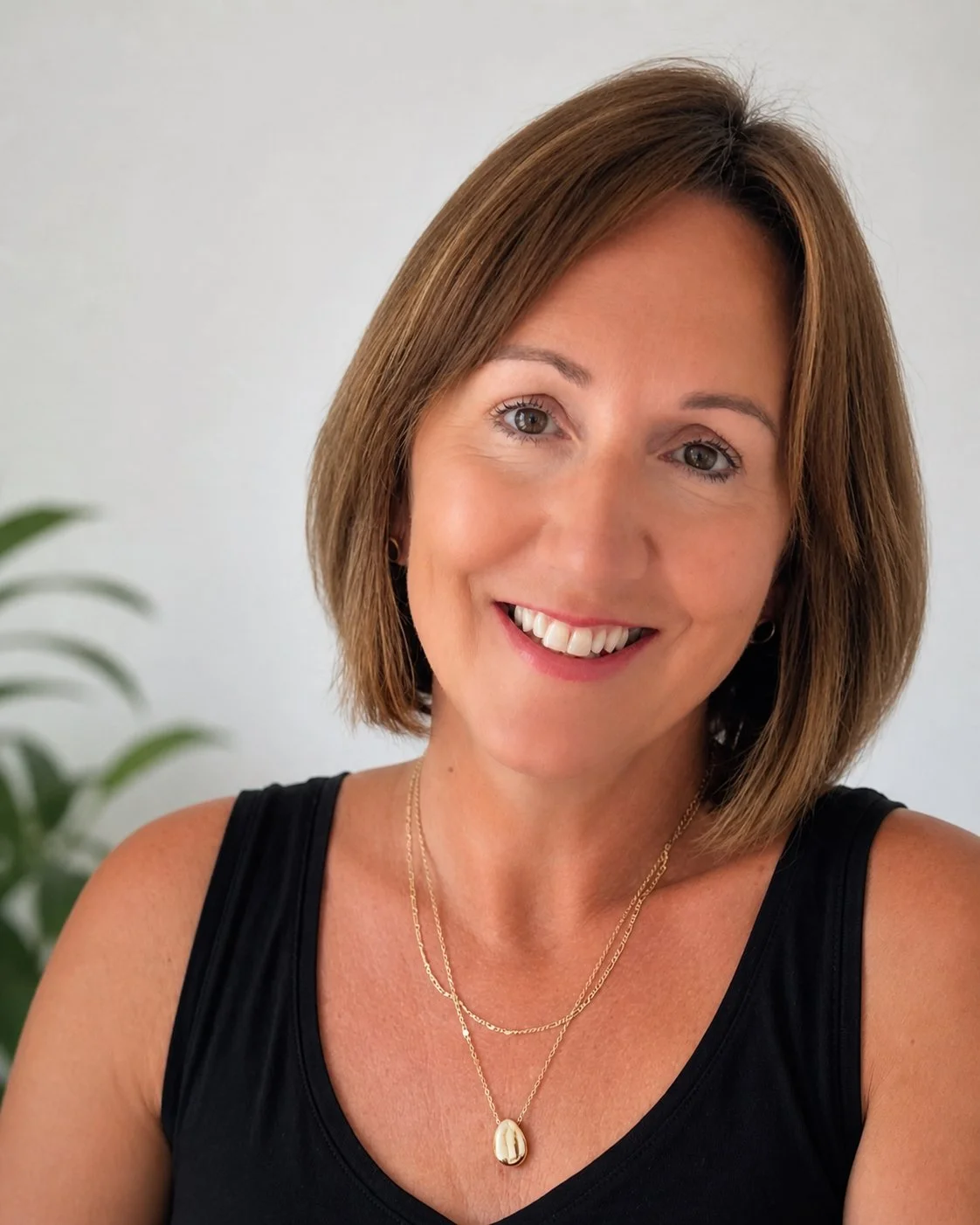 A smiling woman with shoulder-length brown hair wearing a black sleeveless top and layered necklaces, with a light-colored background and a plant in the corner.