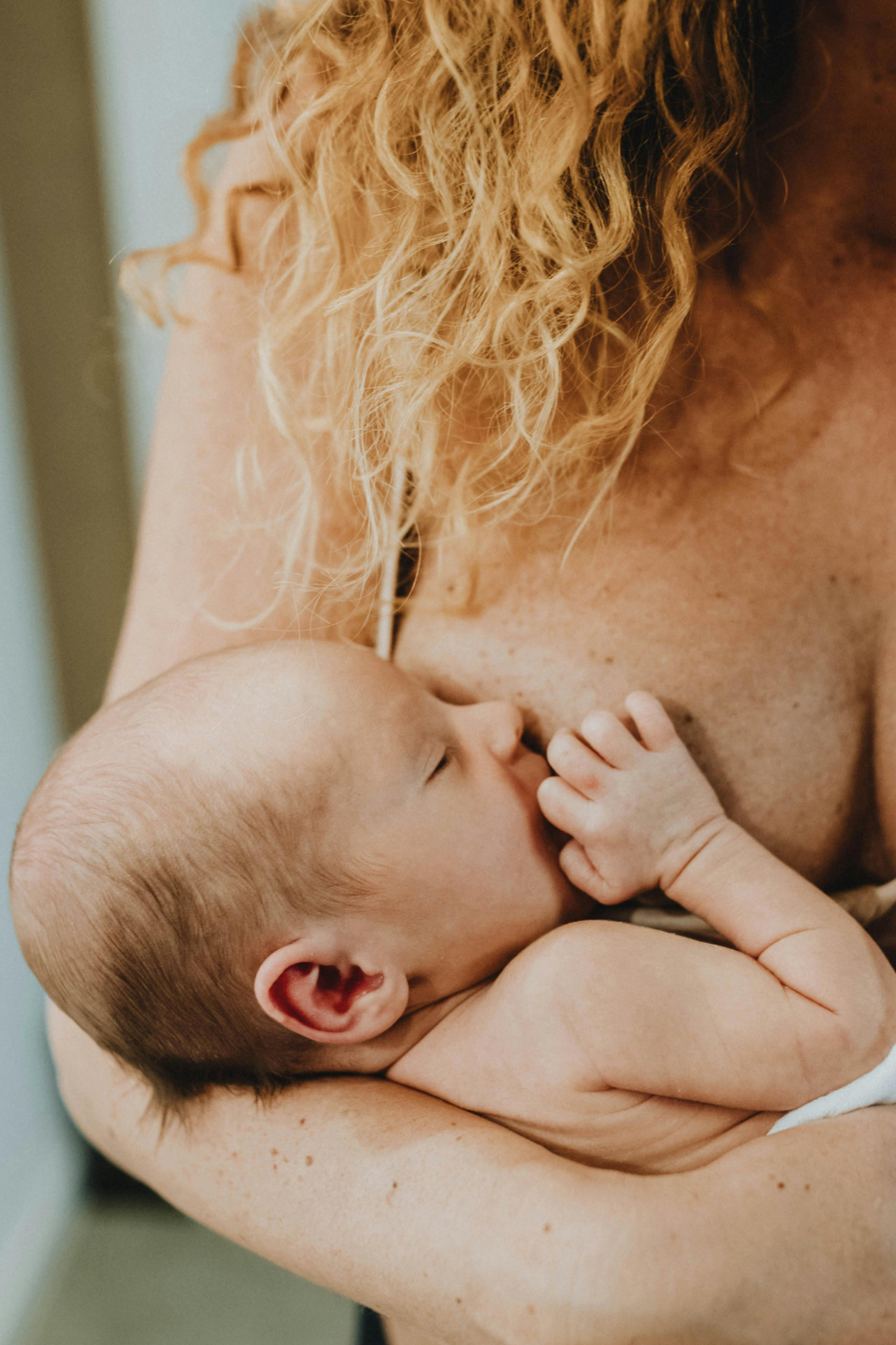 Postpartum mother holding breastfeeding baby while her doula cares for her home and prepares her nourishing food.