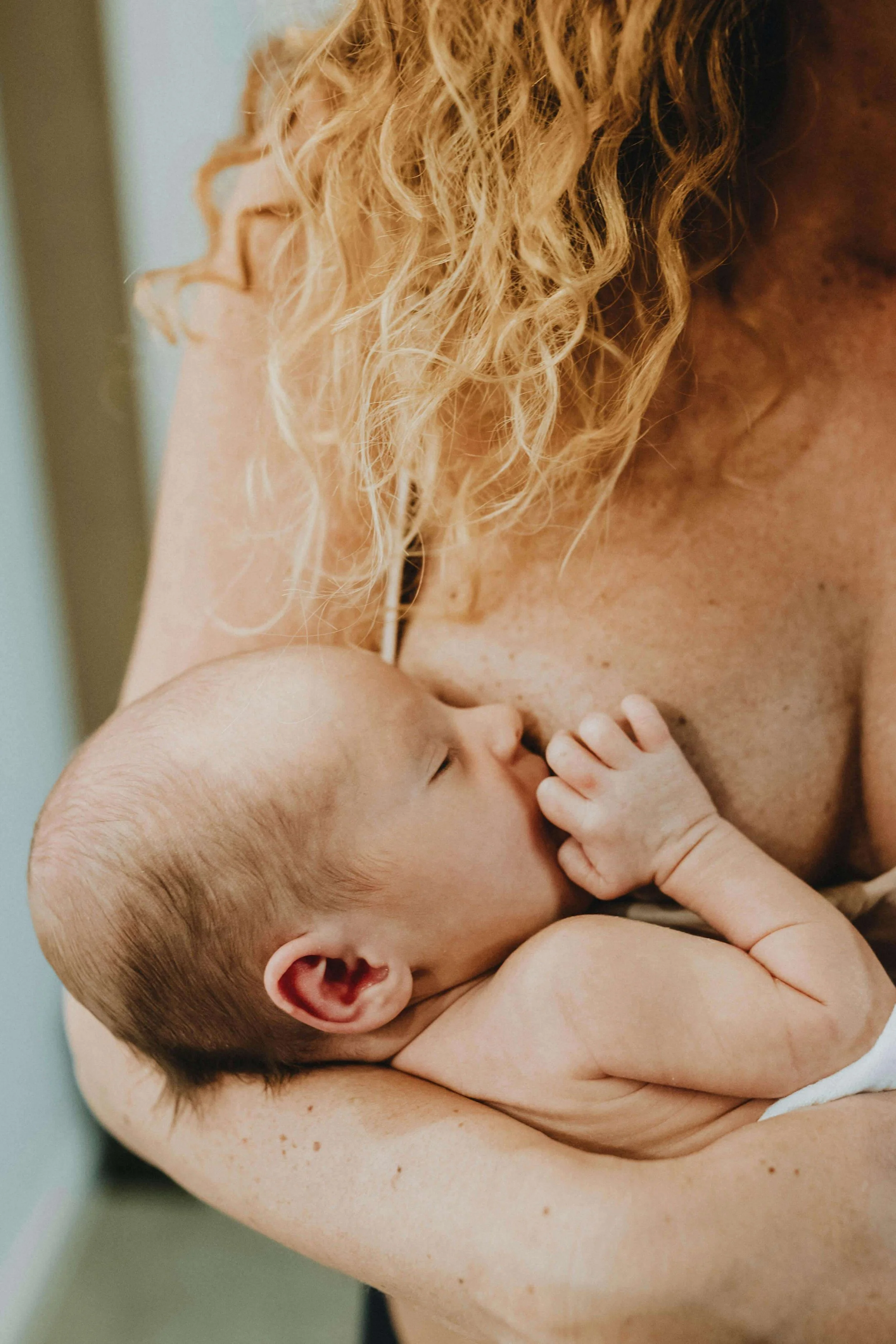 Postpartum mother holding breastfeeding baby while her doula cares for her home and prepares her nourishing food.
