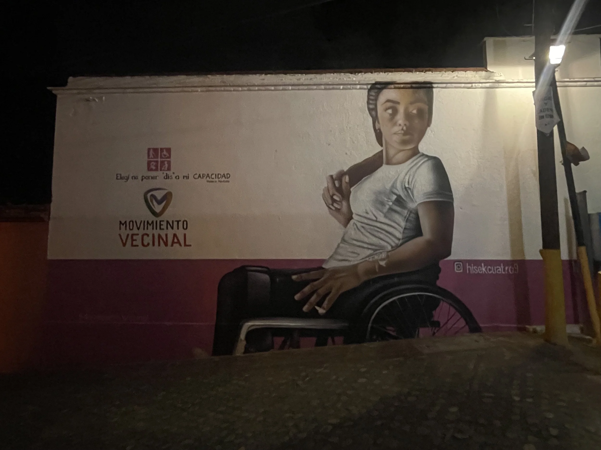 Street mural of a woman with dark hair in a braid, wearing a white t-shirt, using a wheelchair, with text promoting a community movement in Spanish.