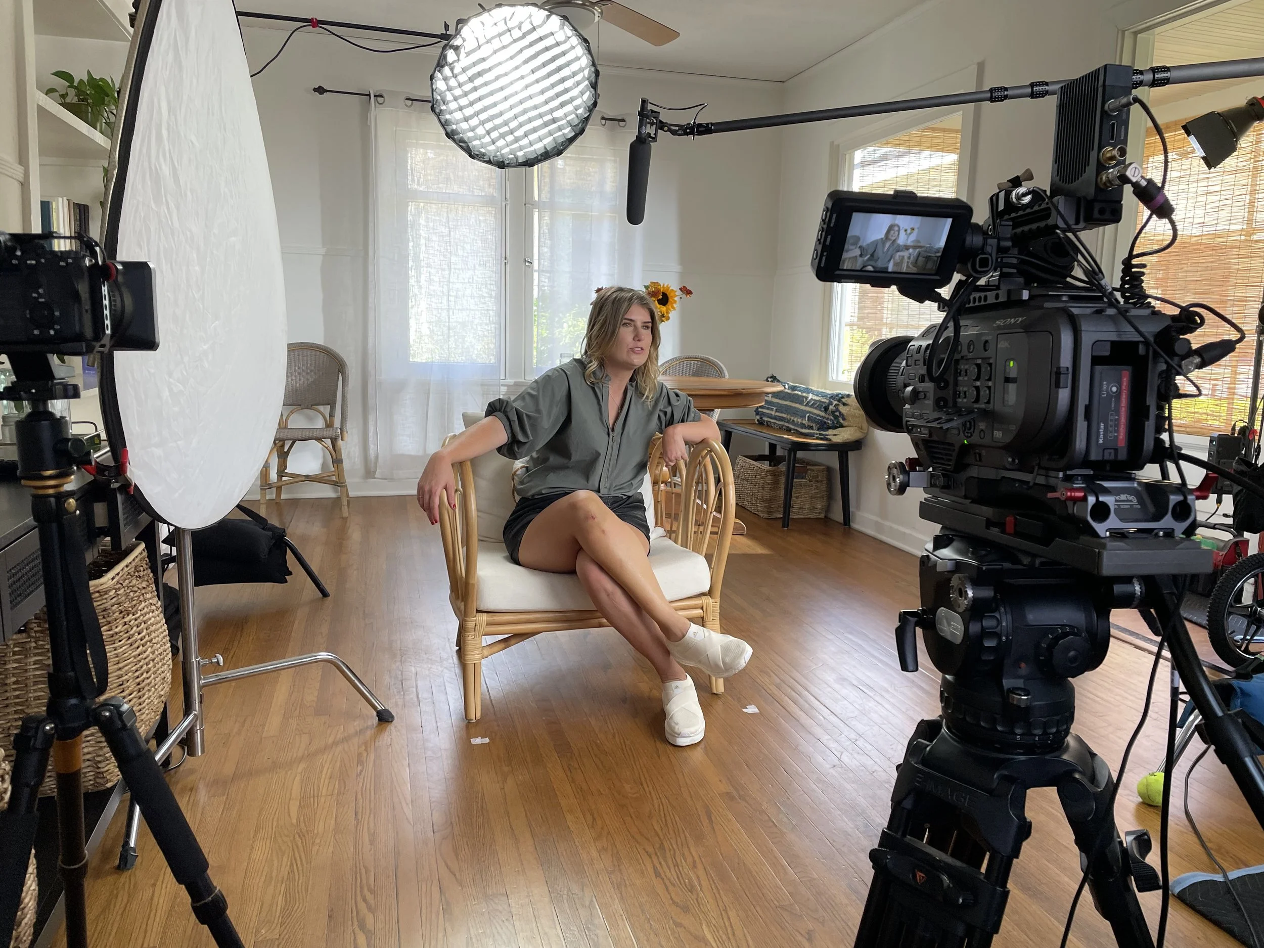 A woman sitting on a chair in a room being filmed for a video interview or production. She is wearing a gray jacket and shorts, with her legs crossed, and is surrounded by professional filming equipment including cameras, lighting, and sound gear. The room has hardwood floors, large windows with blinds, and minimal furniture.