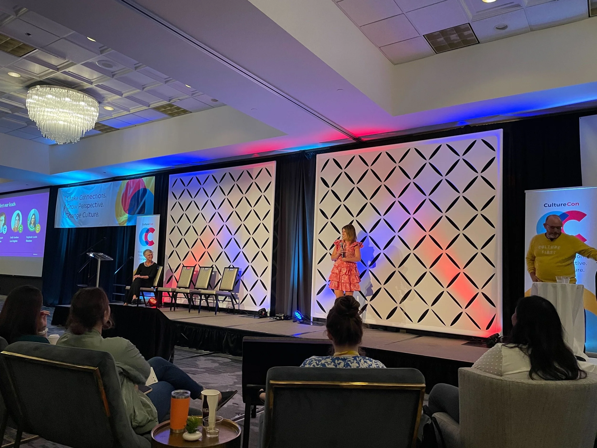 At a conference, a woman in a pink dress speaks at a podium on stage while others sit and stand nearby. Audience members seated in chairs observe the presentation, with some table setups in the foreground. The stage has a decorative backdrop with colorful lighting.