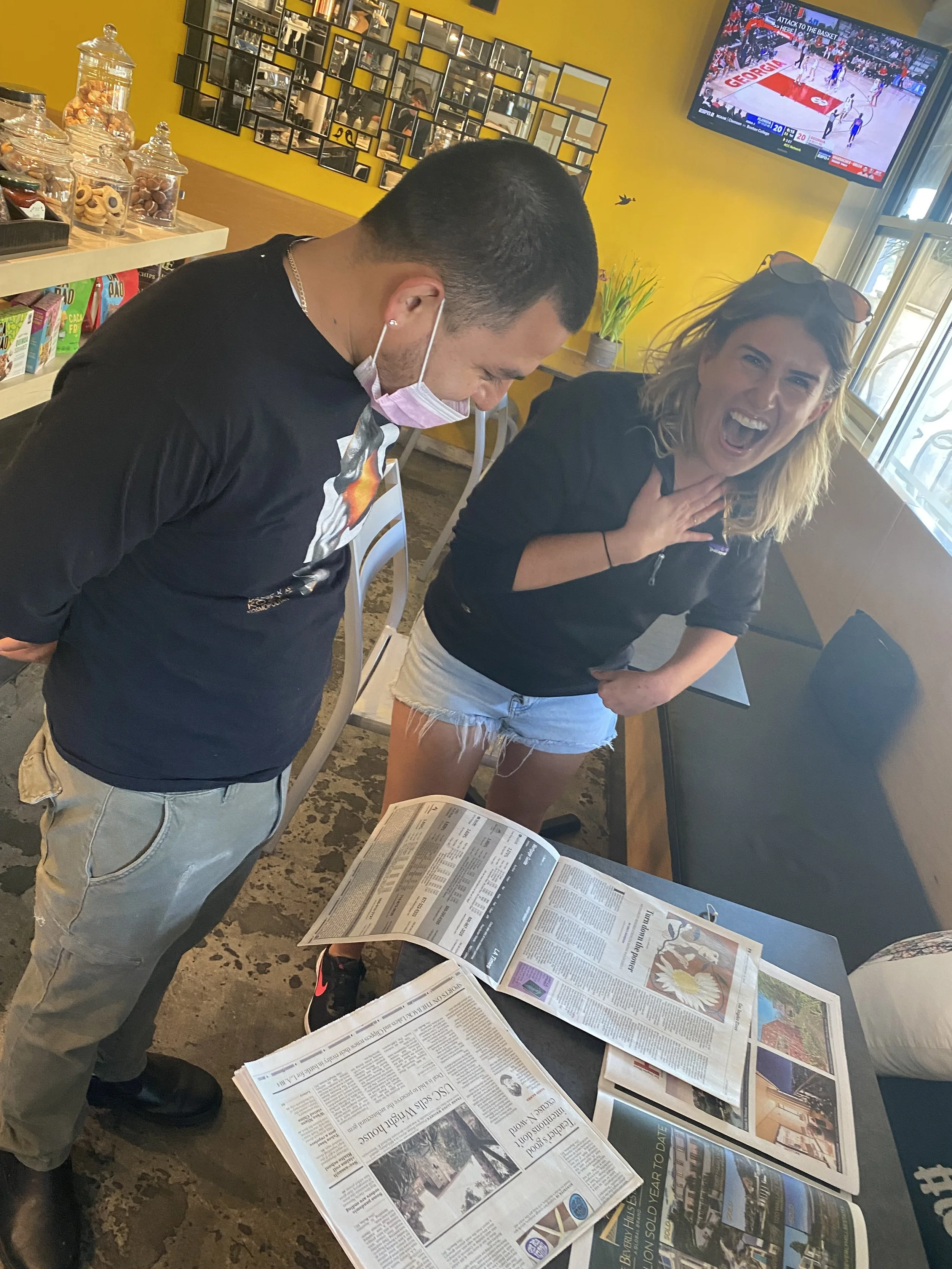 Emily and another person are standing at a table in a cafe looking at her article, "I'm a Disabled Woman. Is That a Dating Dealbreaker?" in the LA Times newspaper. The newspaper is laying on the table in front of them. Emily is laughing.
