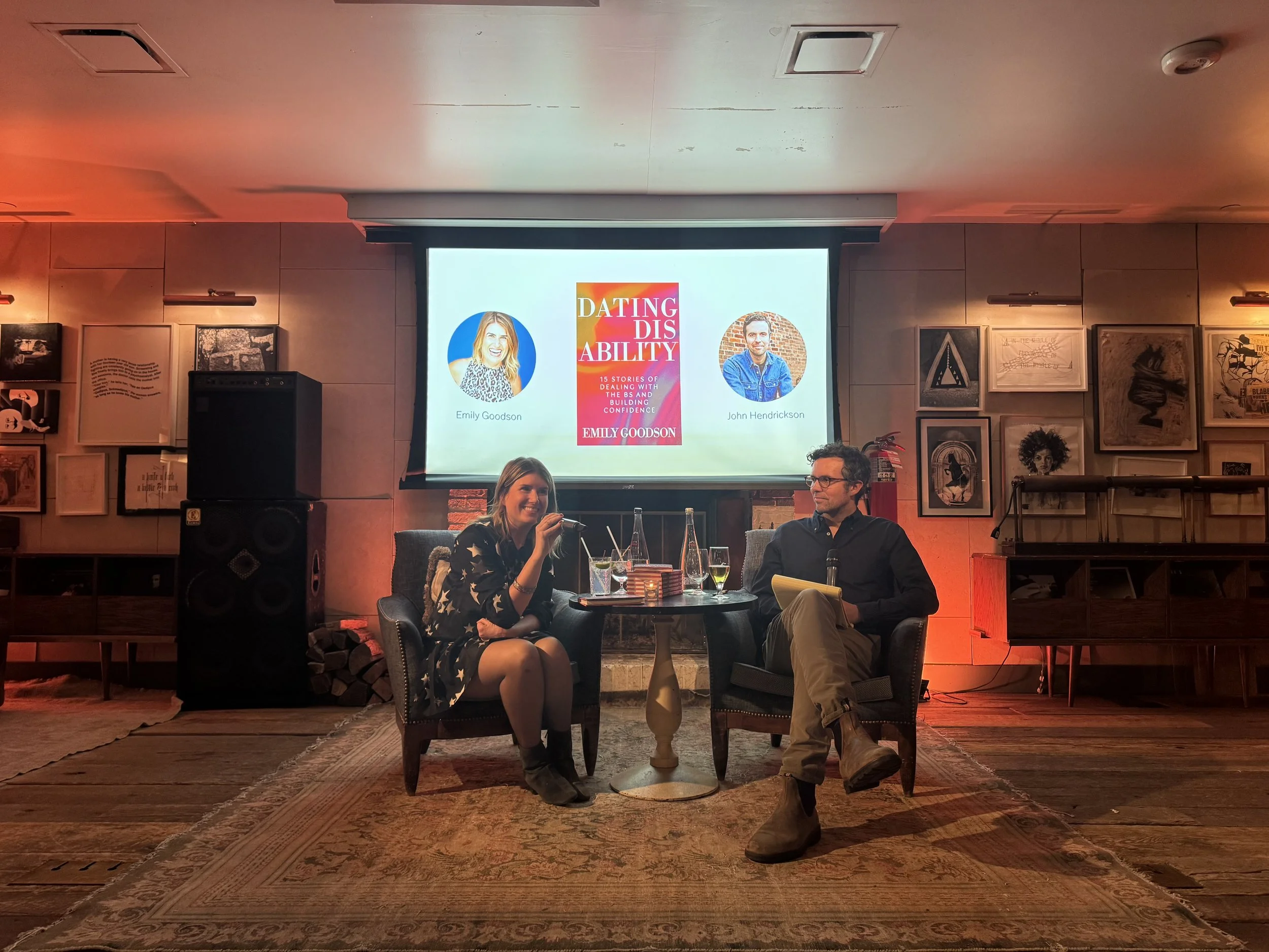Two people sitting on chairs in front of a large screen discussing a book titled 'Dating Disability' in a cozy, art-filled room with framed pictures on the walls and a carpeted floor.