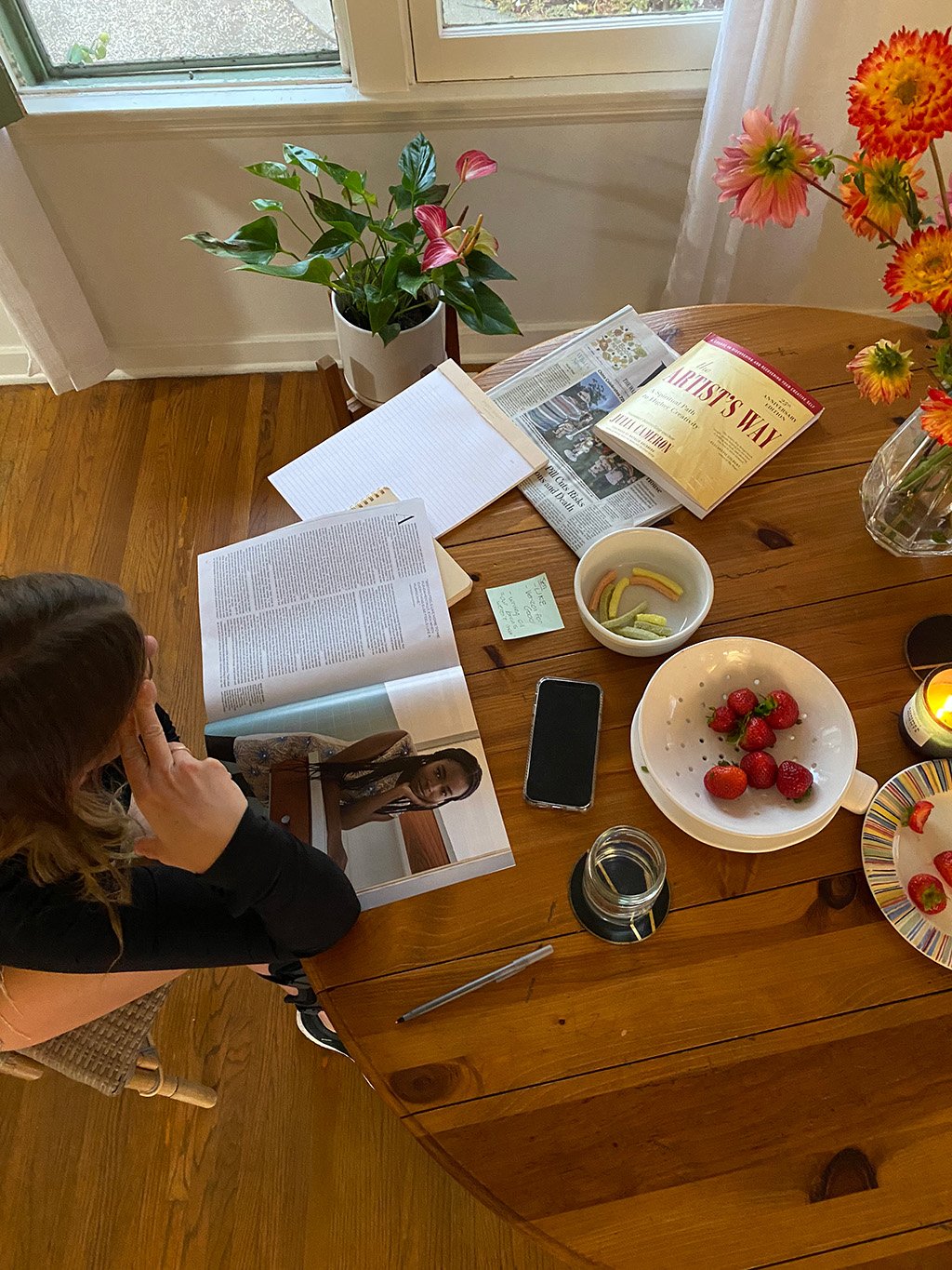 A wooden dining table with various items including books, newspapers, a bowl of strawberries, a plate of strawberries on a colorful plate, a glass jar, a smartphone, a pen, and a small note. There are also flowers in a vase and a potted plant nearby.