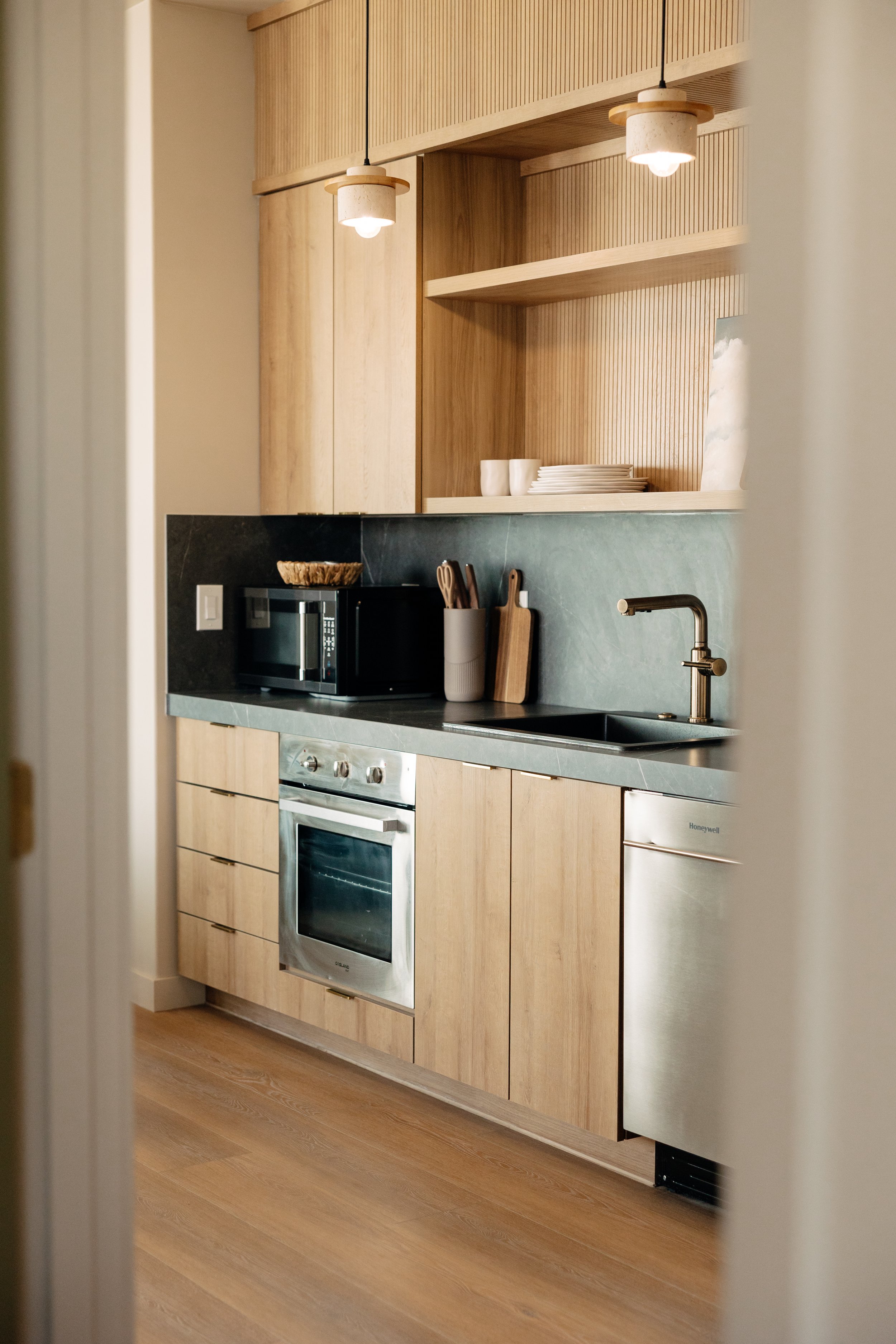 A modern kitchen with light wood cabinets, a black countertop, a stainless steel oven, and stainless steel appliances. There are open shelves with dishes, a microwave, and some utensils on the counter. Two pendant lights hang from the ceiling, and th