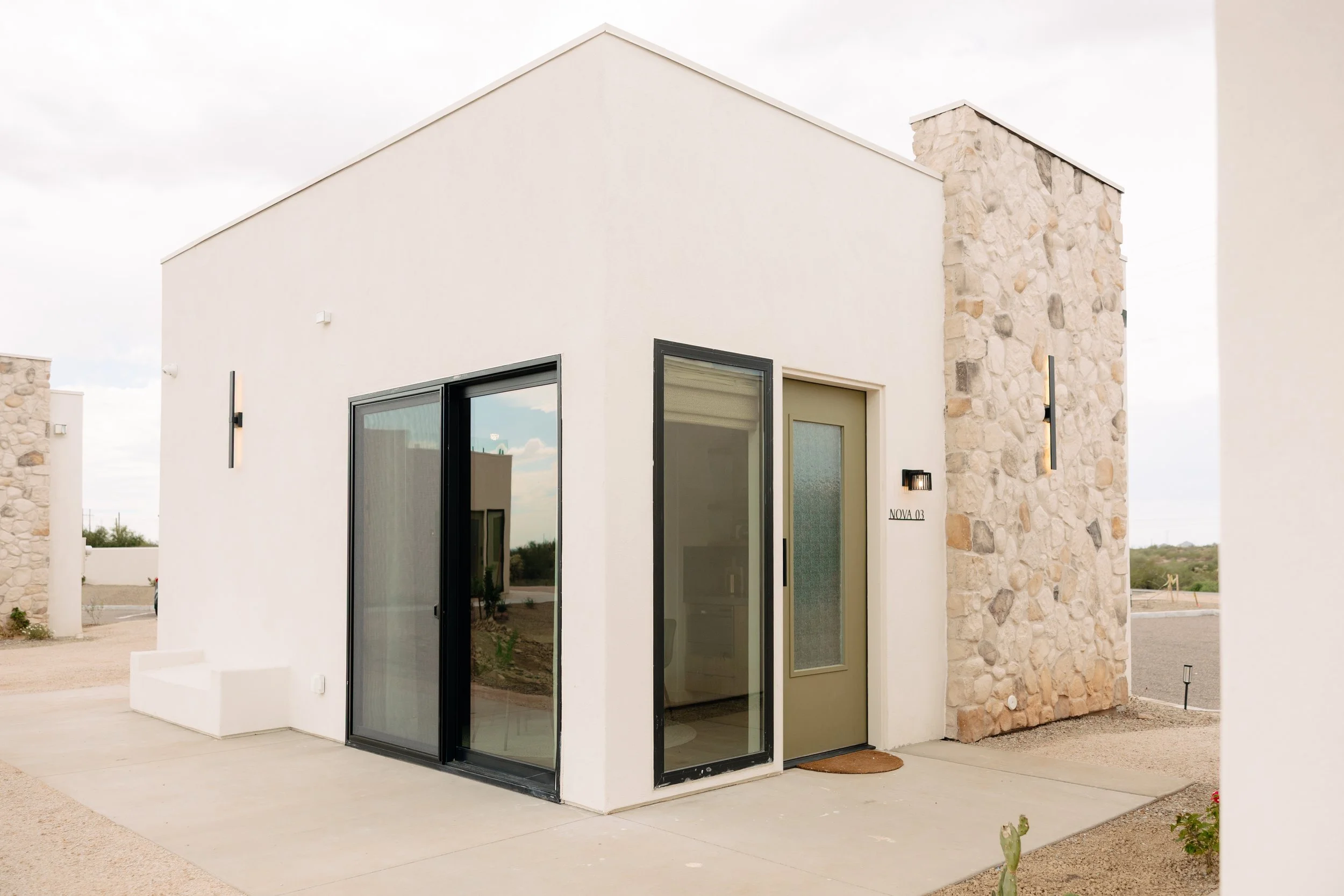Modern small white house with stone accents, glass sliding doors, and an olive-green front door, situated in a desert landscape.