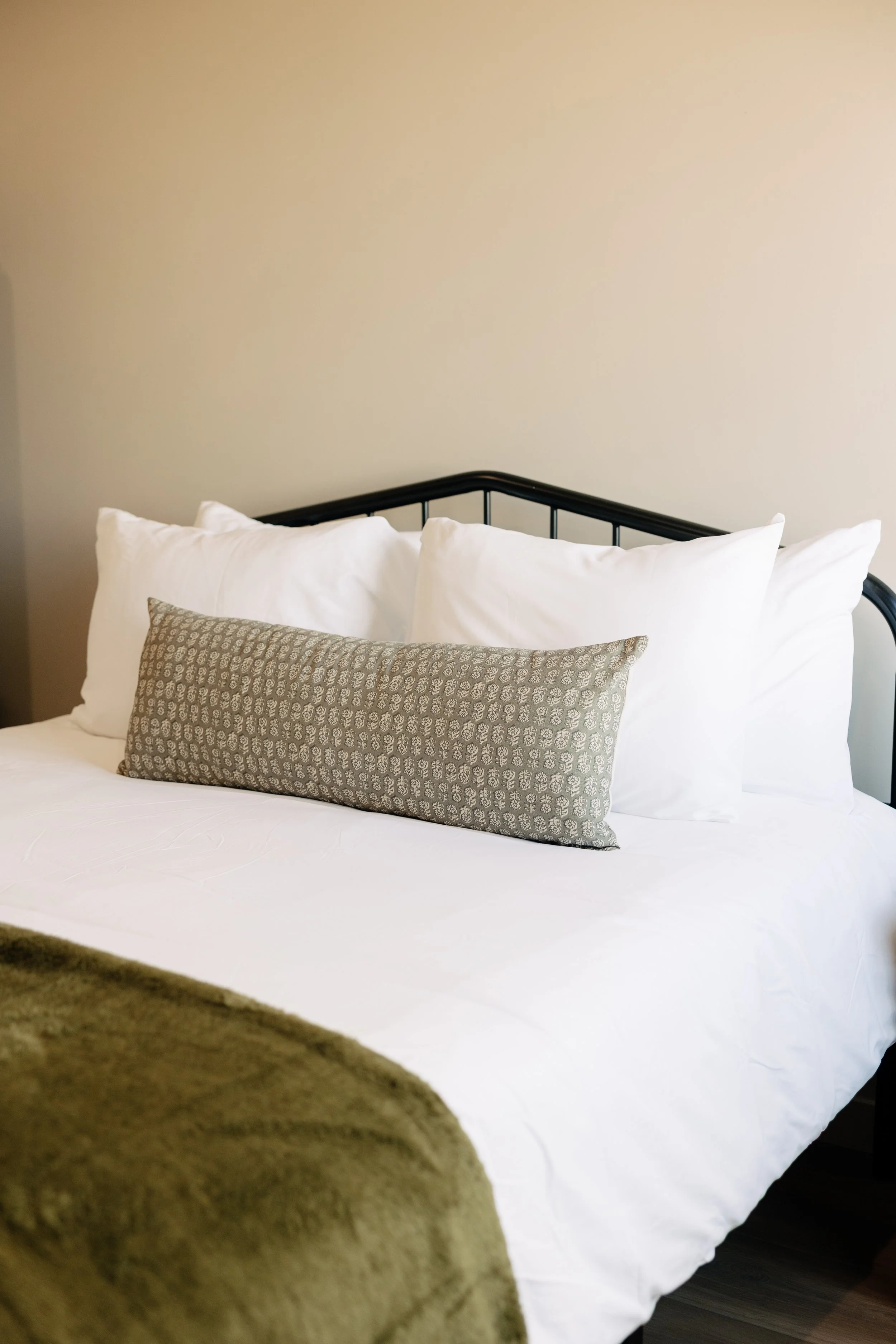 A neatly made bed with white sheets, four white pillows, a gray decorative pillow, and a green bedspread in a hotel room with beige walls.