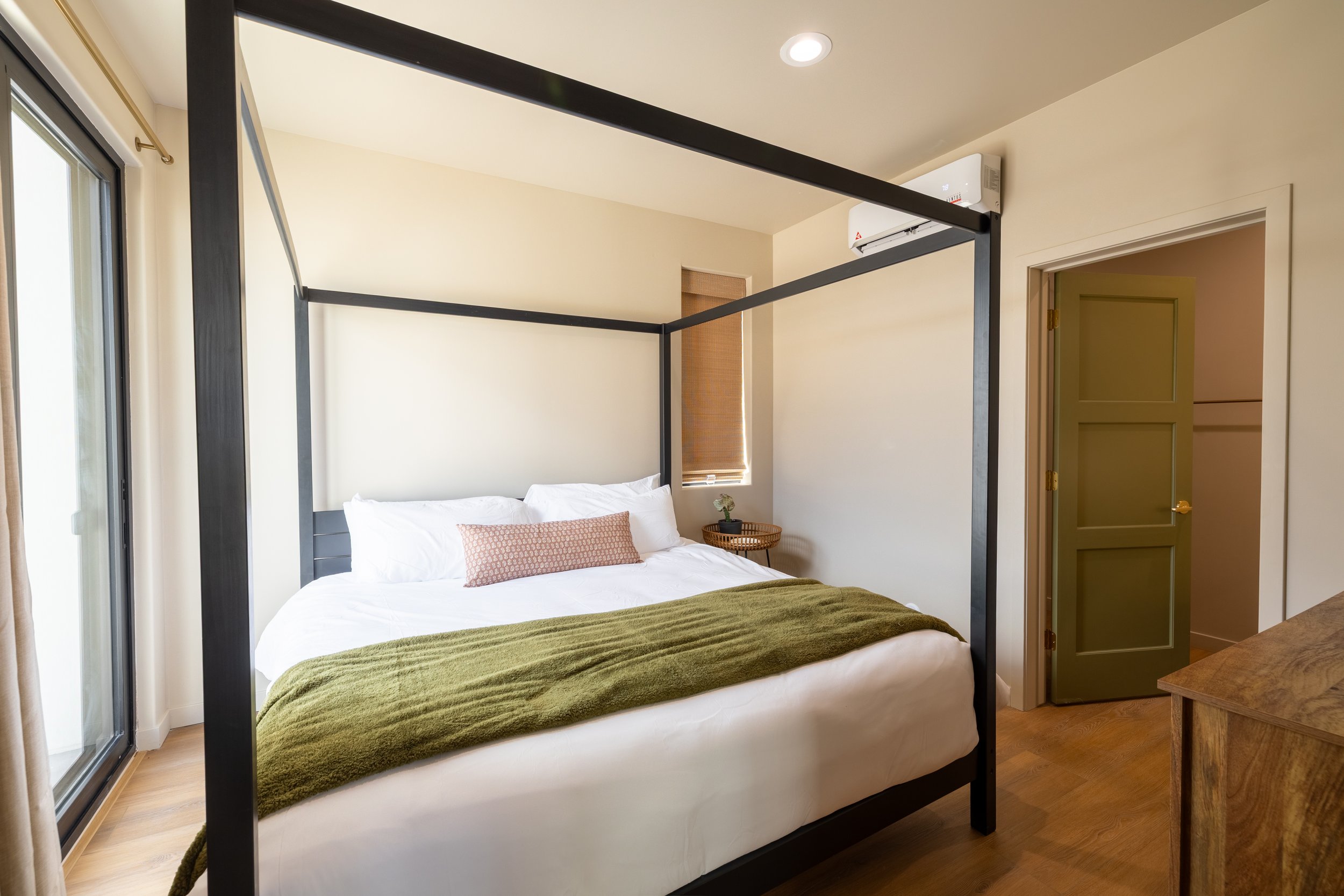 A bedroom with a black four-poster bed, white bedding, an olive green throw, and a decorative pillow. There is a small window with a brown shade, a side table with a small plant, and a sliding glass door leading outside. An air conditioning unit is m