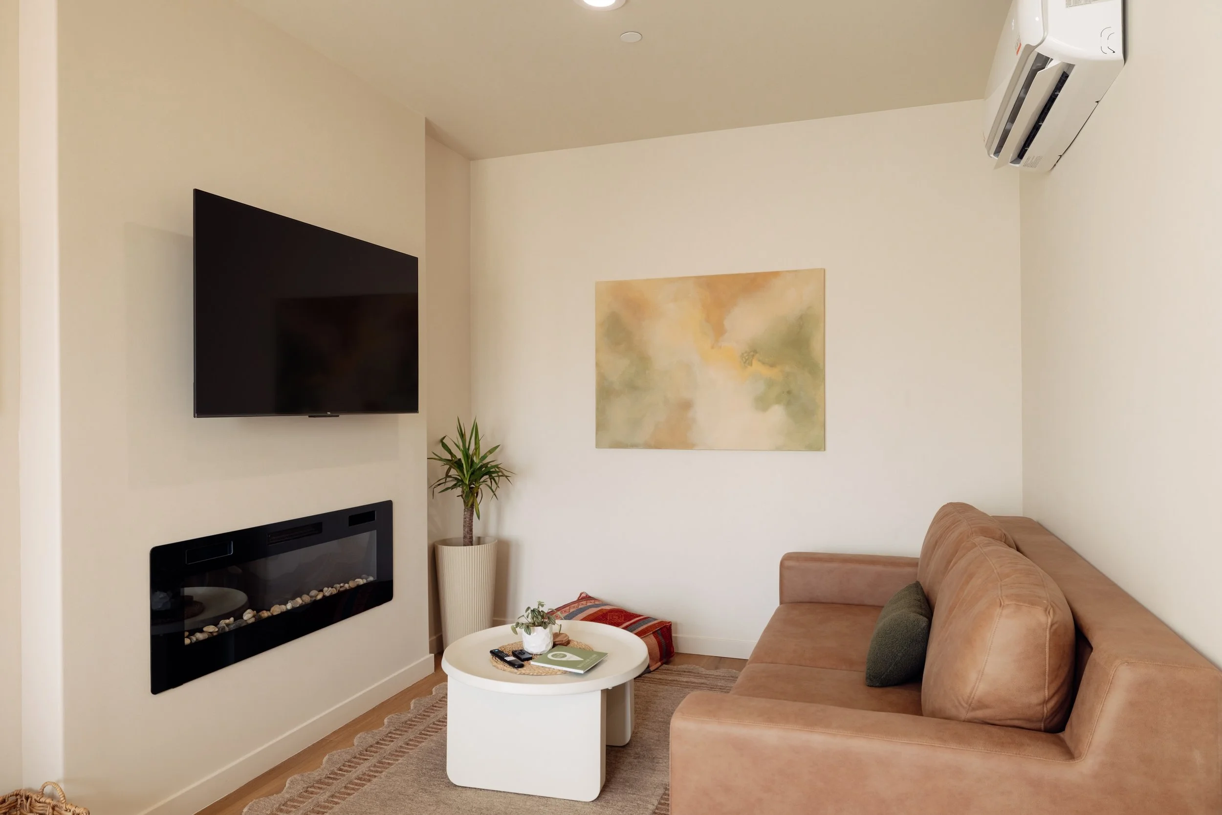 Cozy living room with a wall-mounted flat-screen TV and an electric fireplace beneath it, a tan leather sofa with a green pillow, a small white coffee table with remote controls and a plant, a painted artwork on the wall, a potted plant in a tall bei