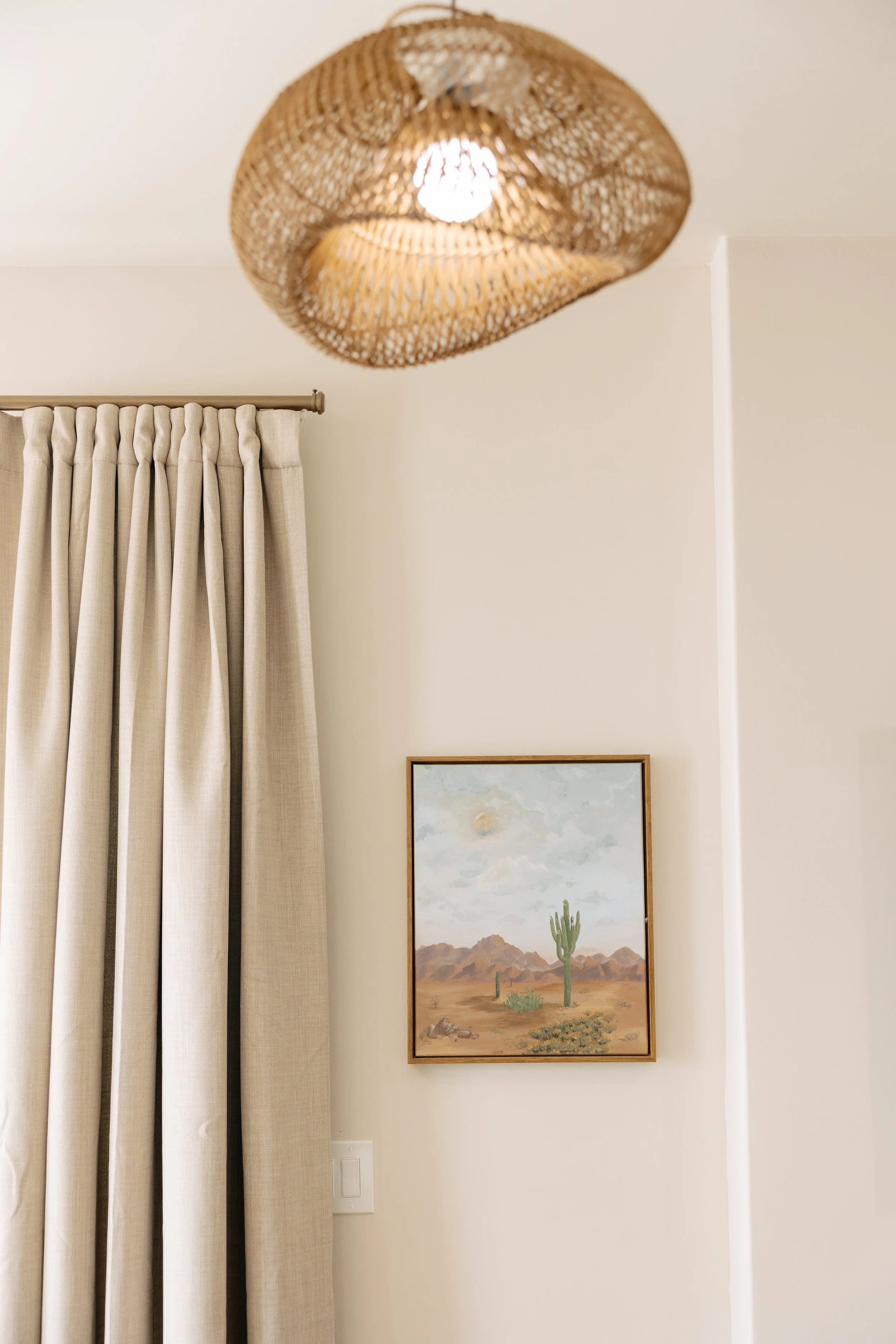 Interior of a room with beige curtains, a framed desert landscape painting with cacti and mountains, and a wicker ceiling light