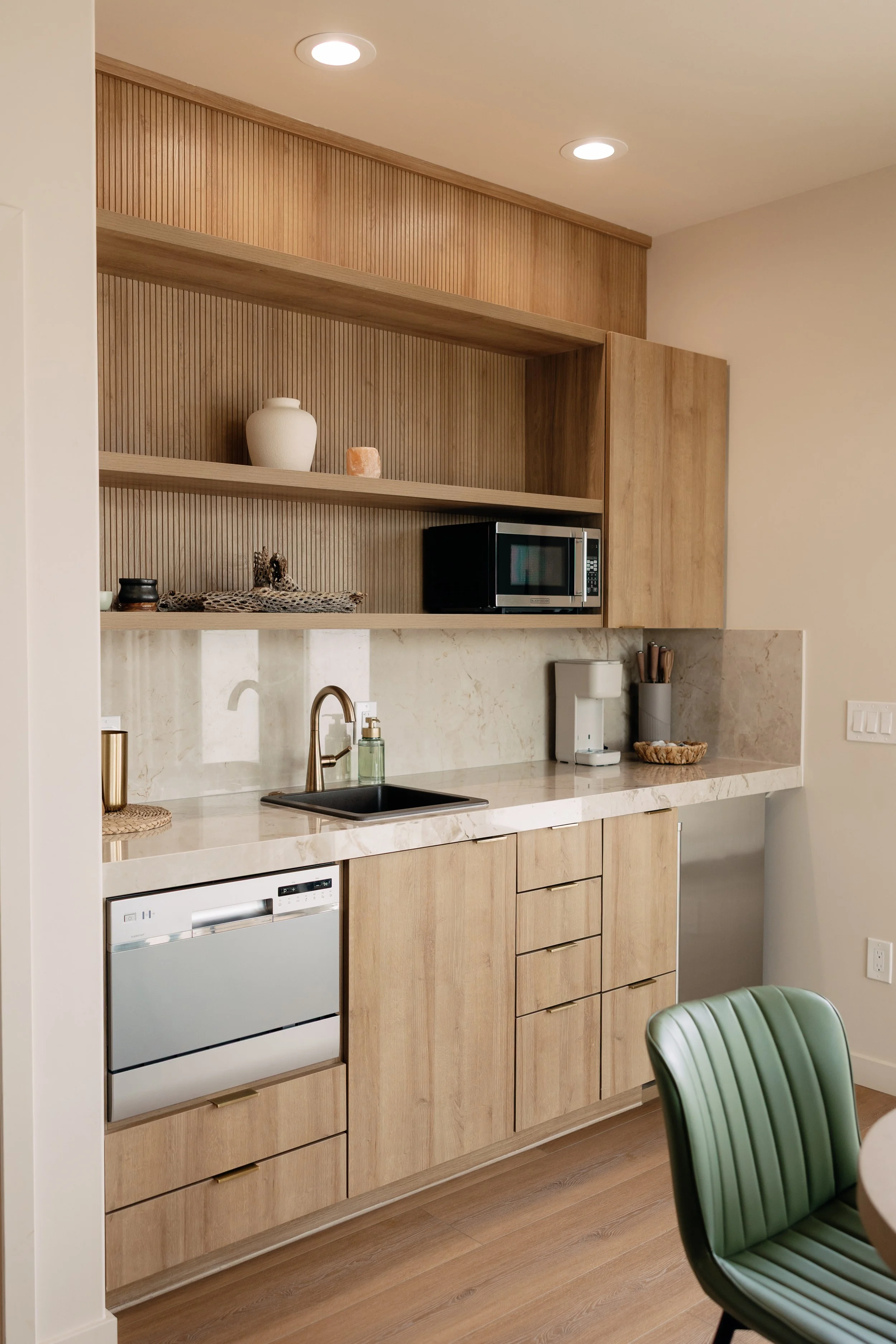 Modern kitchen with light wood cabinetry, marble countertop, small sink, microwave, coffee maker, decorative items, green chair, and warm lighting.