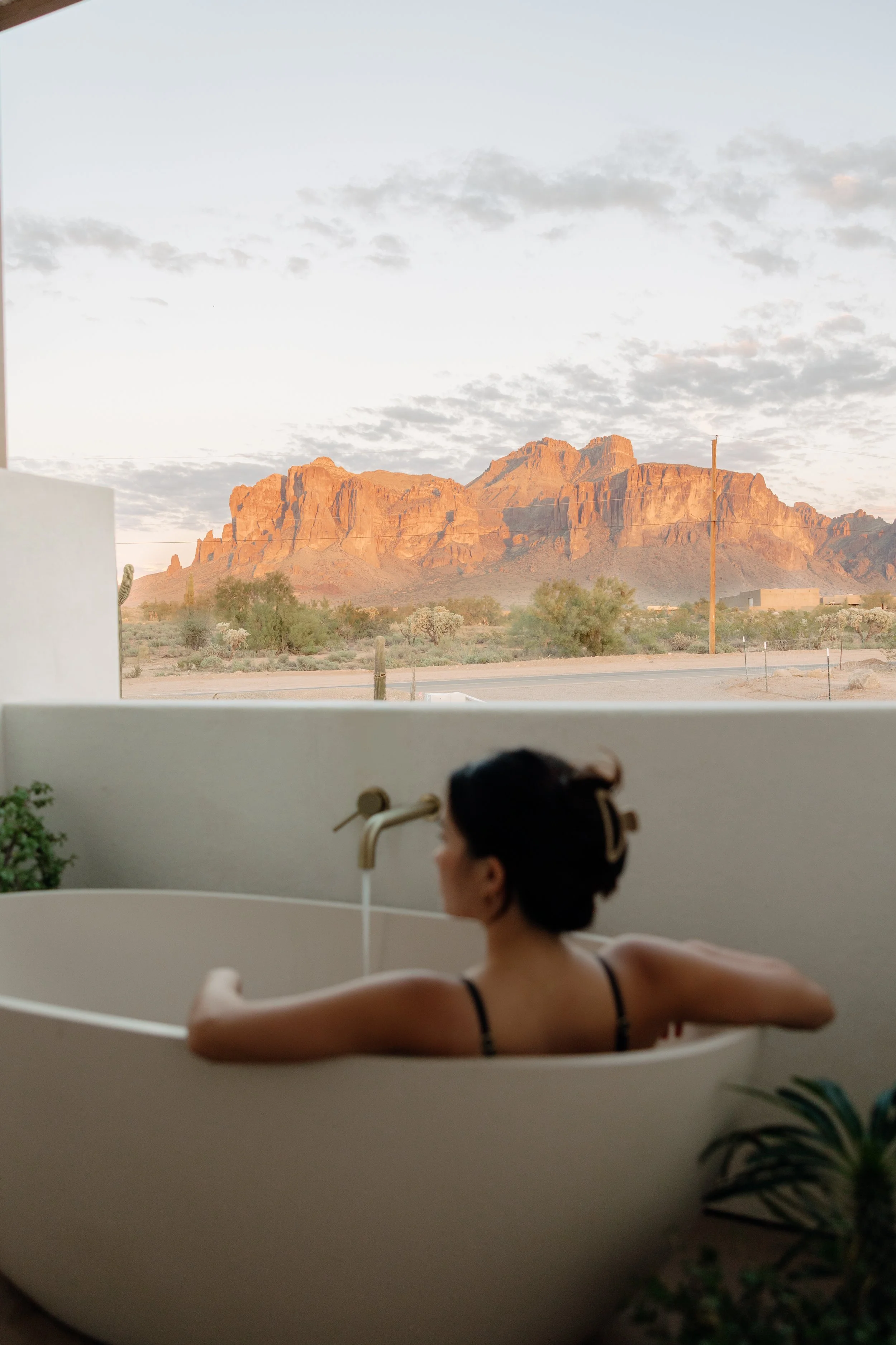 A woman relaxing in an outdoor bathtub, overlooking a desert landscape with mountains in the distance during sunset.