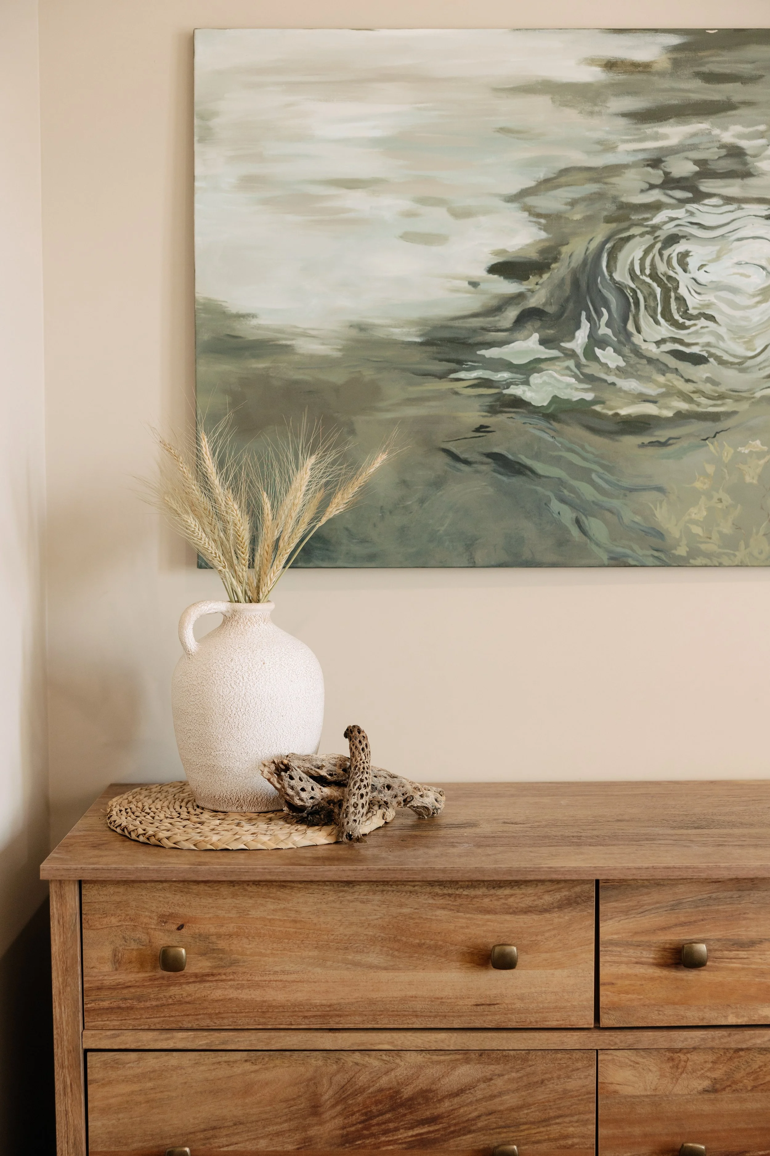 A wooden dresser with a white textured vase containing dried grass, and decorative driftwood pieces on top, with an abstract ocean painting hanging on the wall behind.