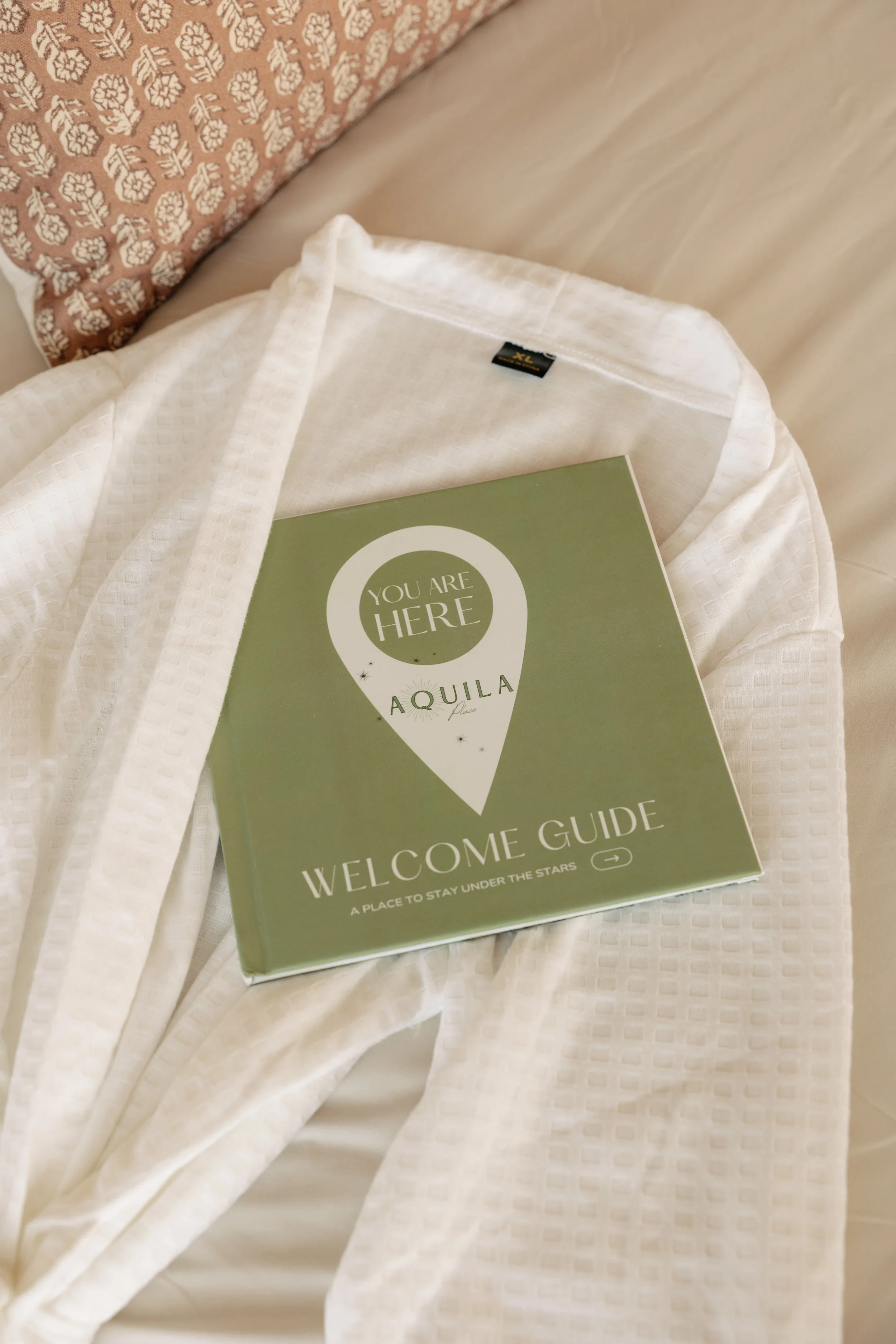 A welcome guide booklet titled 'You Are Here Aquila Place' resting on a white robe or towel, with a pillow featuring a patterned pillowcase in the background.