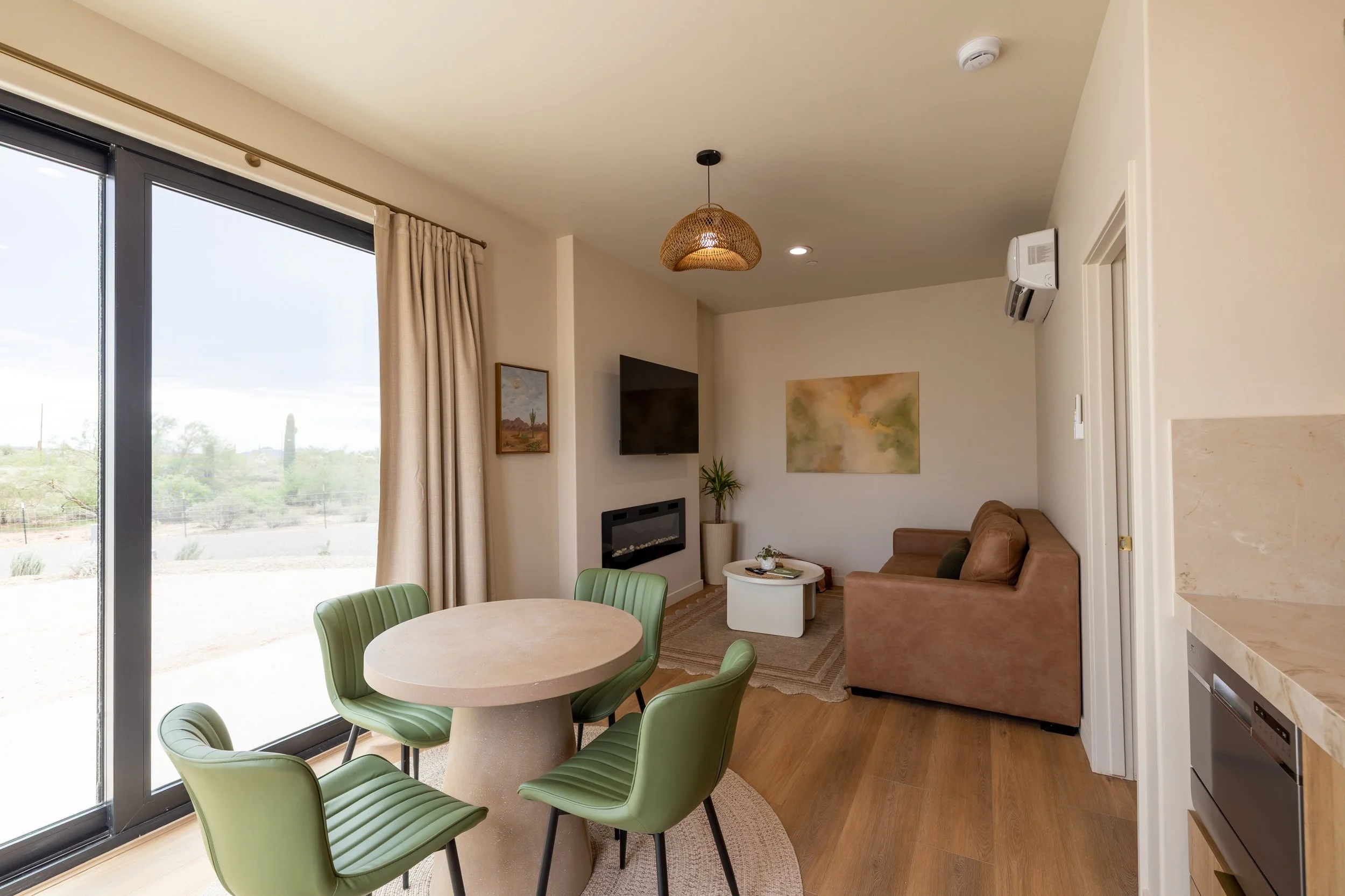 Modern living room with a round dining table and six green chairs, beige curtains, a brown sofa, a wall-mounted TV, abstract wall art, a fireplace, a potted plant, and a sliding glass door showing a desert landscape.