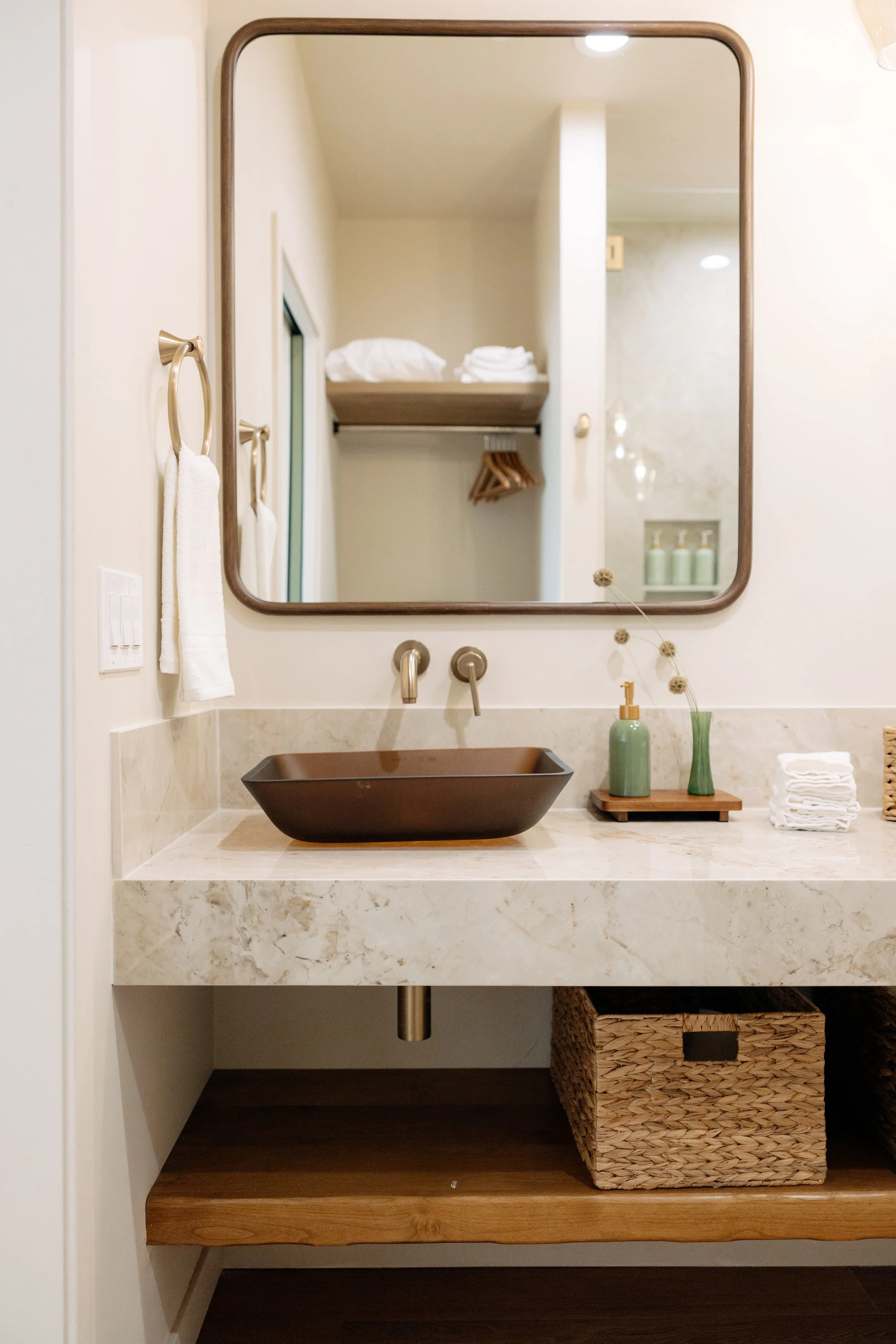 Modern bathroom vanity with a brown vessel sink, wall-mounted faucet, large mirror, hand towels, decorative bottles, and woven storage baskets.