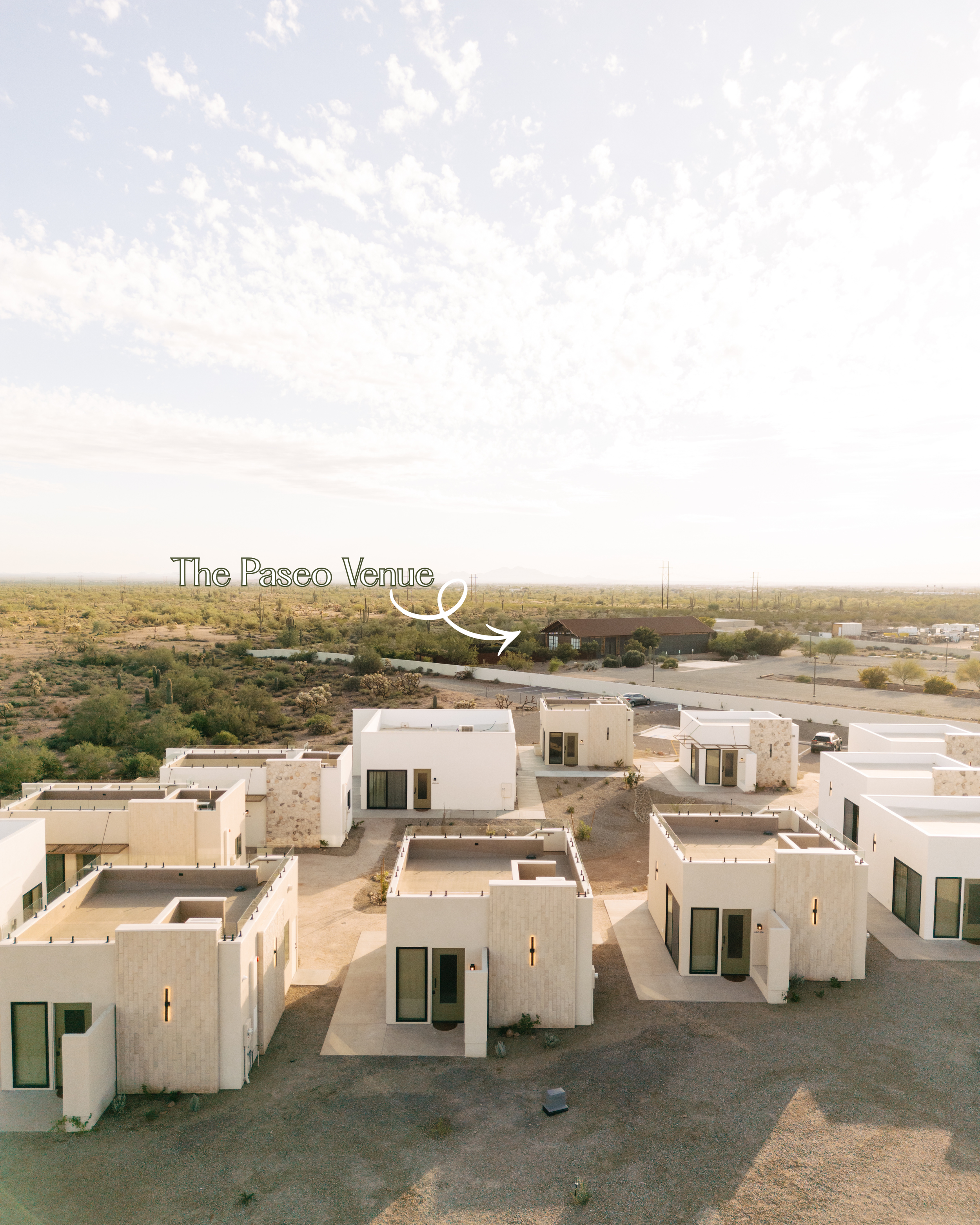 Aerial view of modern white houses with flat roofs in a desert landscape, with a sign pointing to 'The Paseo Venue' in the distance.