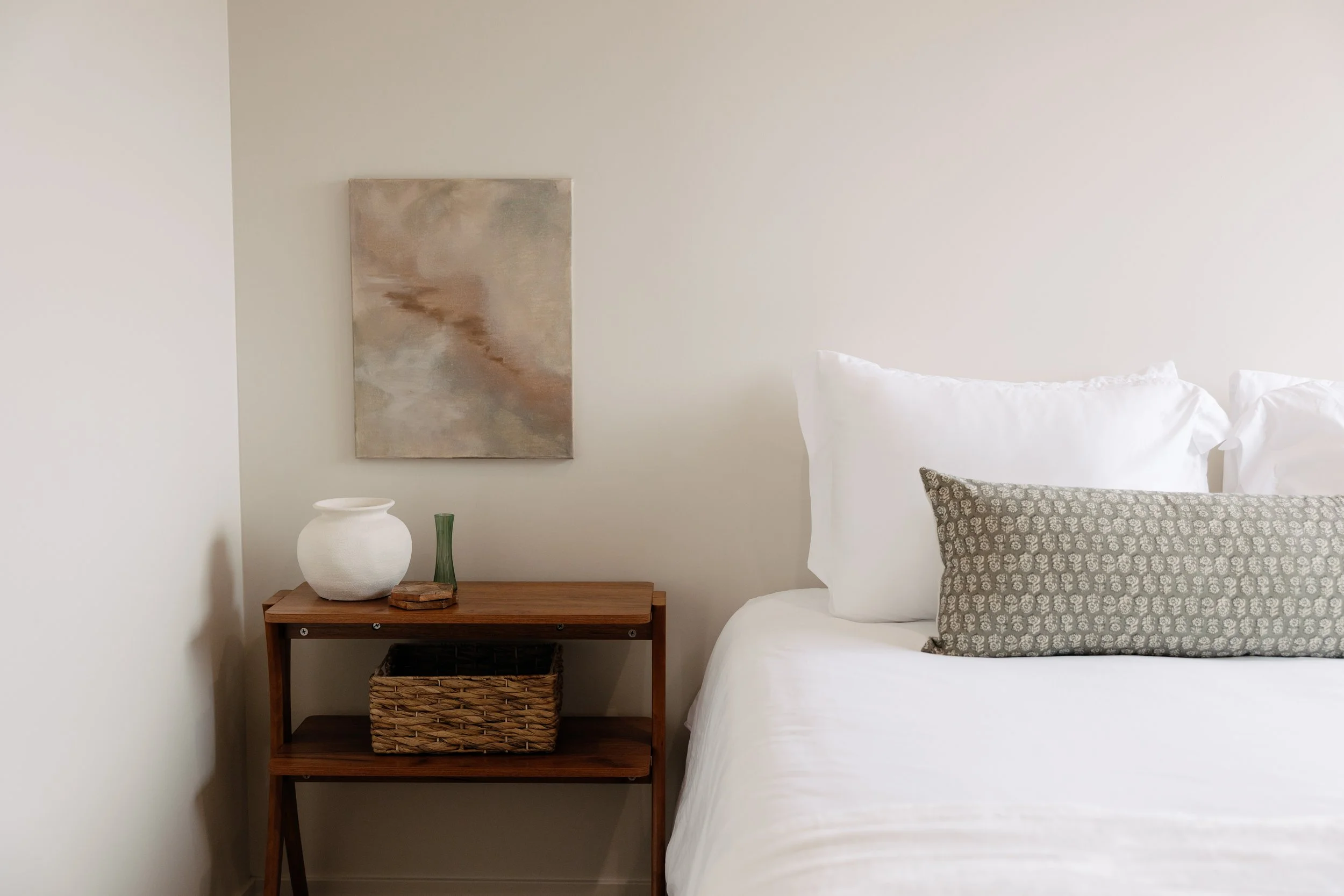 A bedroom with a bed, white linens, and decorative pillows. Next to the bed, a wooden nightstand holds a white vase, a green vase, presents, and a wicker basket underneath. A framed abstract painting hangs on the wall above the nightstand.
