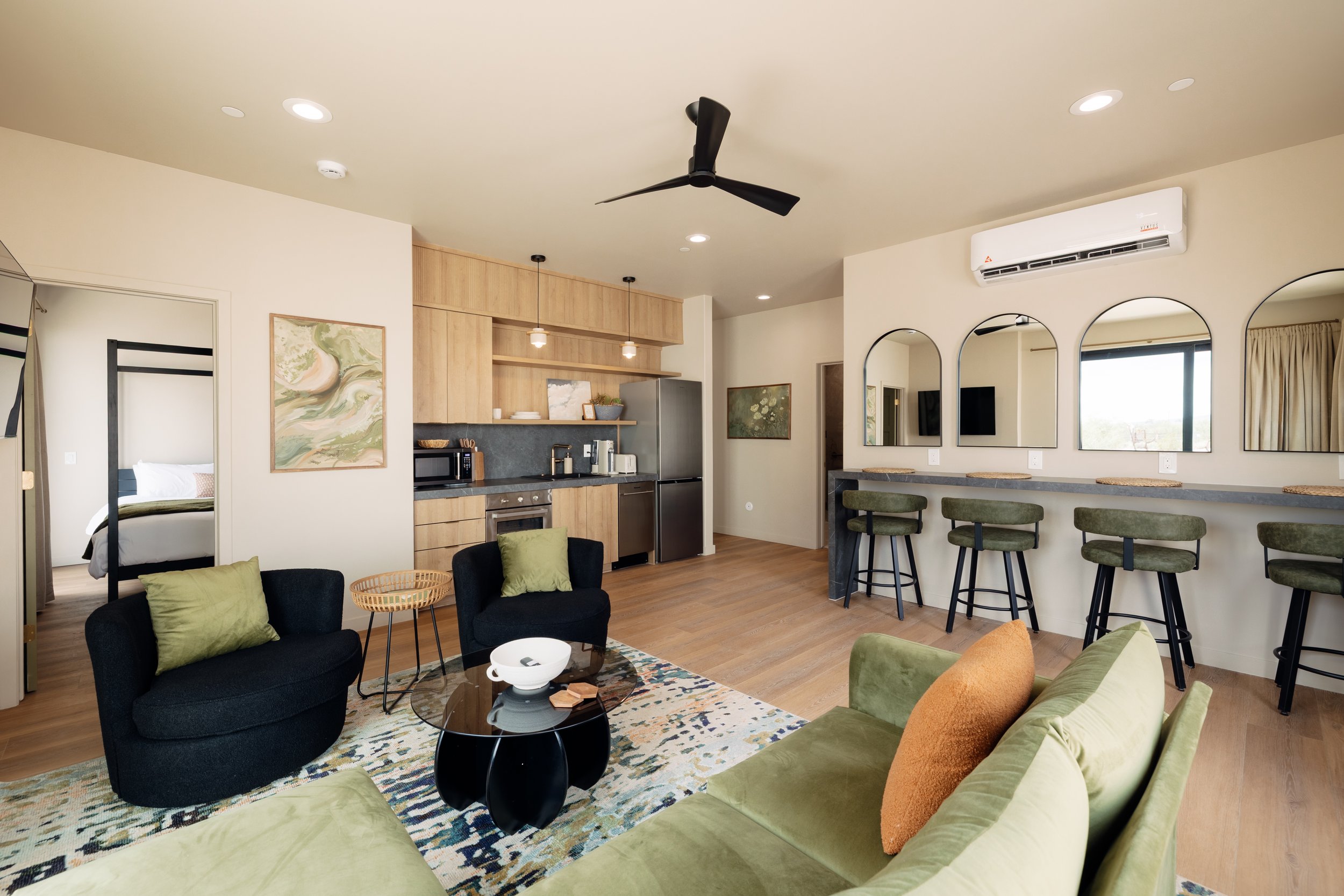 Living room with black and green sofas, a glass coffee table, a kitchen area with wooden cabinets, a bar with four stools, a large window, and a bedroom visible through an open doorway.