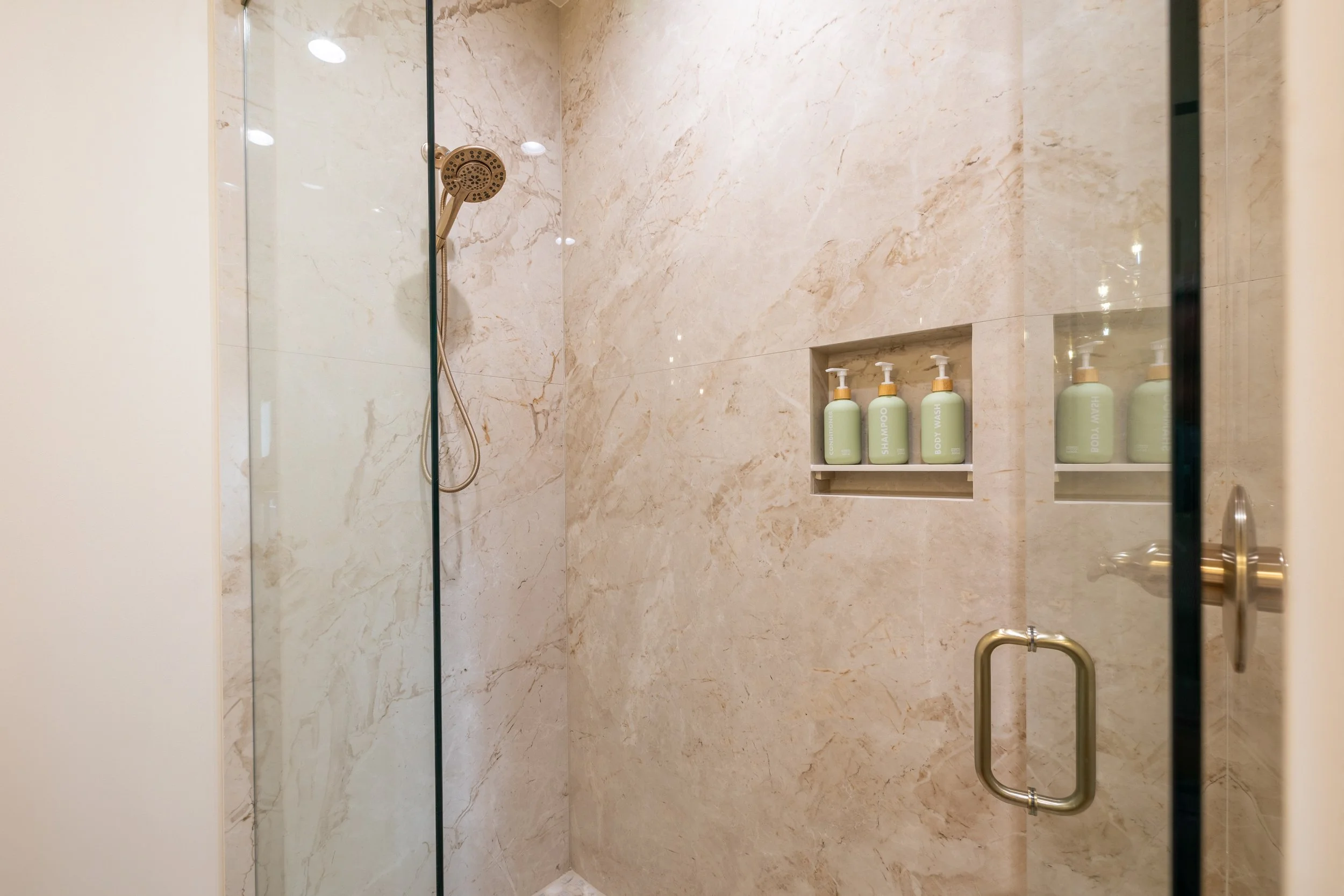 A modern shower with beige marble walls and a glass door. There is a built-in shelf with three bottles of body wash, shampoo, and lotion. The shower has a round, wall-mounted showerhead and a handheld shower.