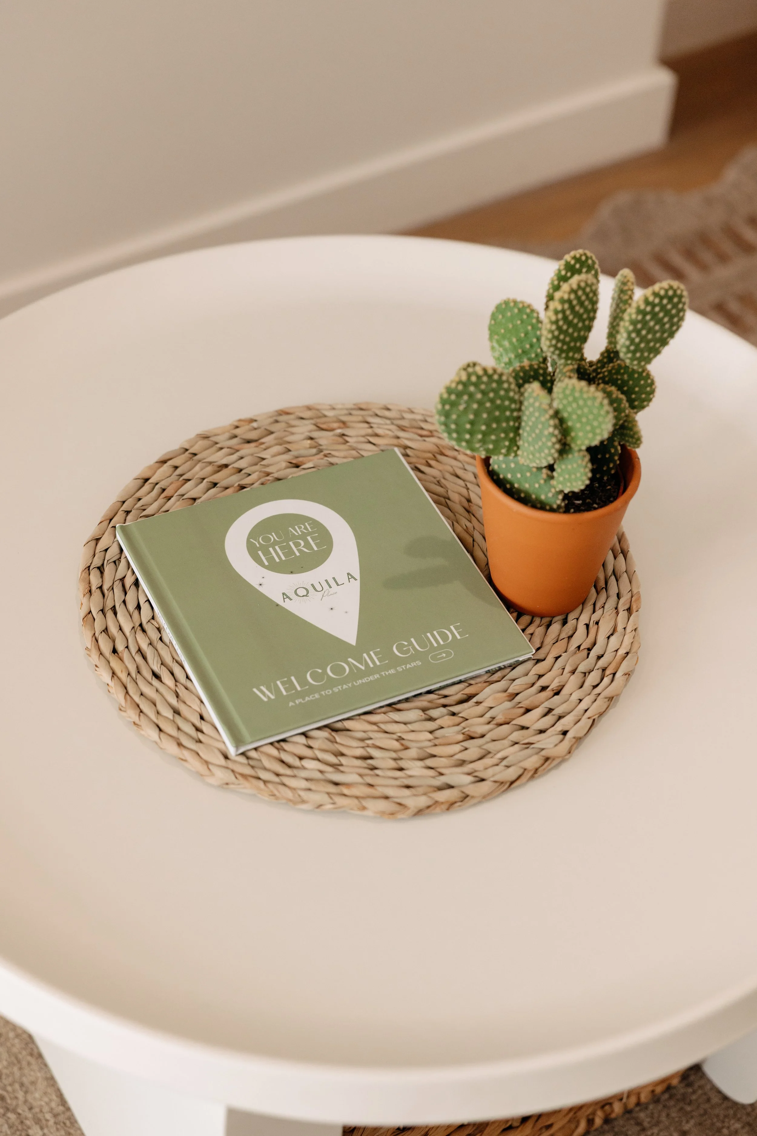A round white table with a woven placemat, a potted cactus, and a green welcome guide booklet.