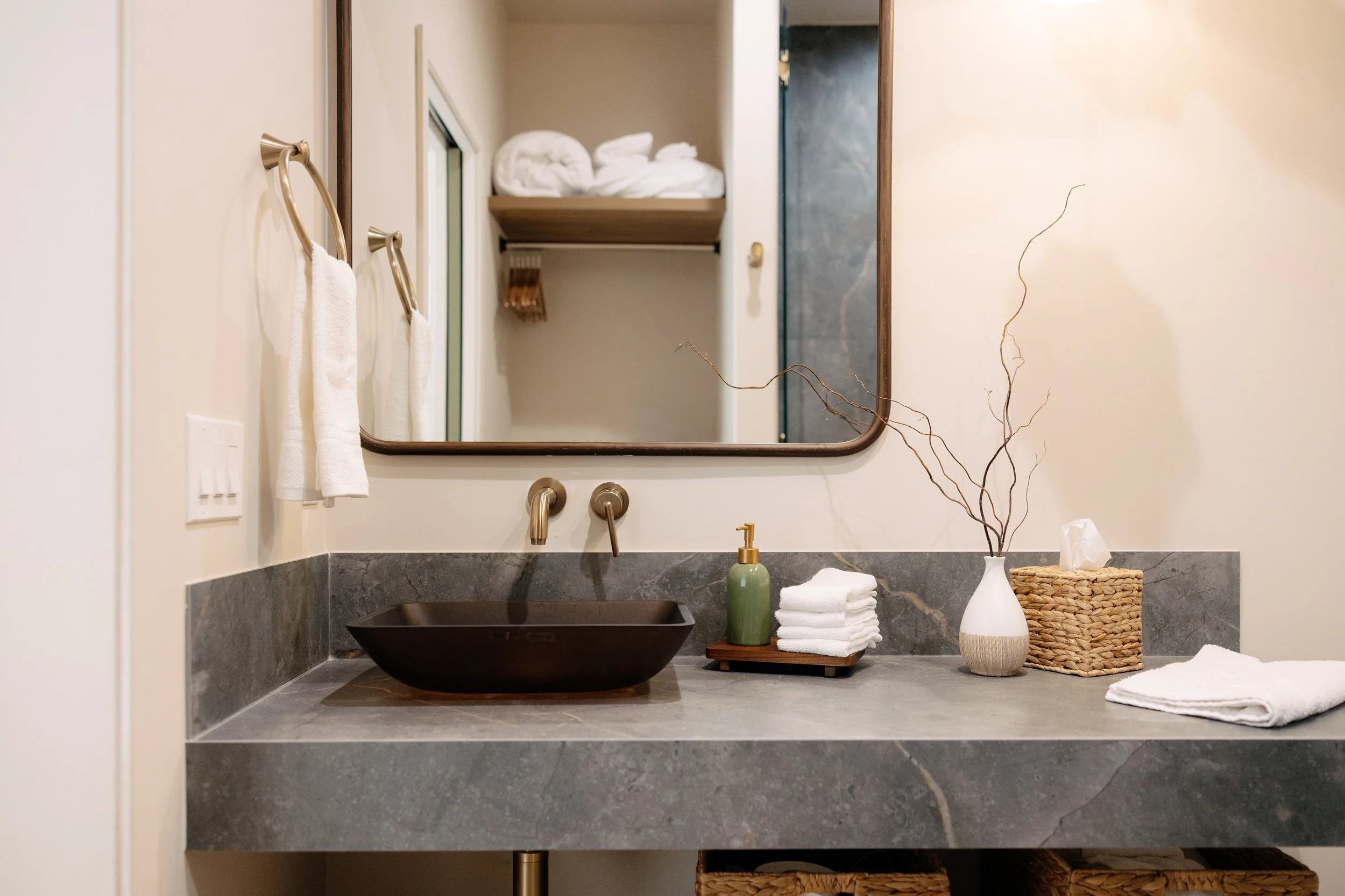 Modern bathroom vanity with a dark brown vessel sink, gold faucet, mirror, and decor including a vase with dry branches, neatly folded white towels, a tissue box, and a wicker basket.
