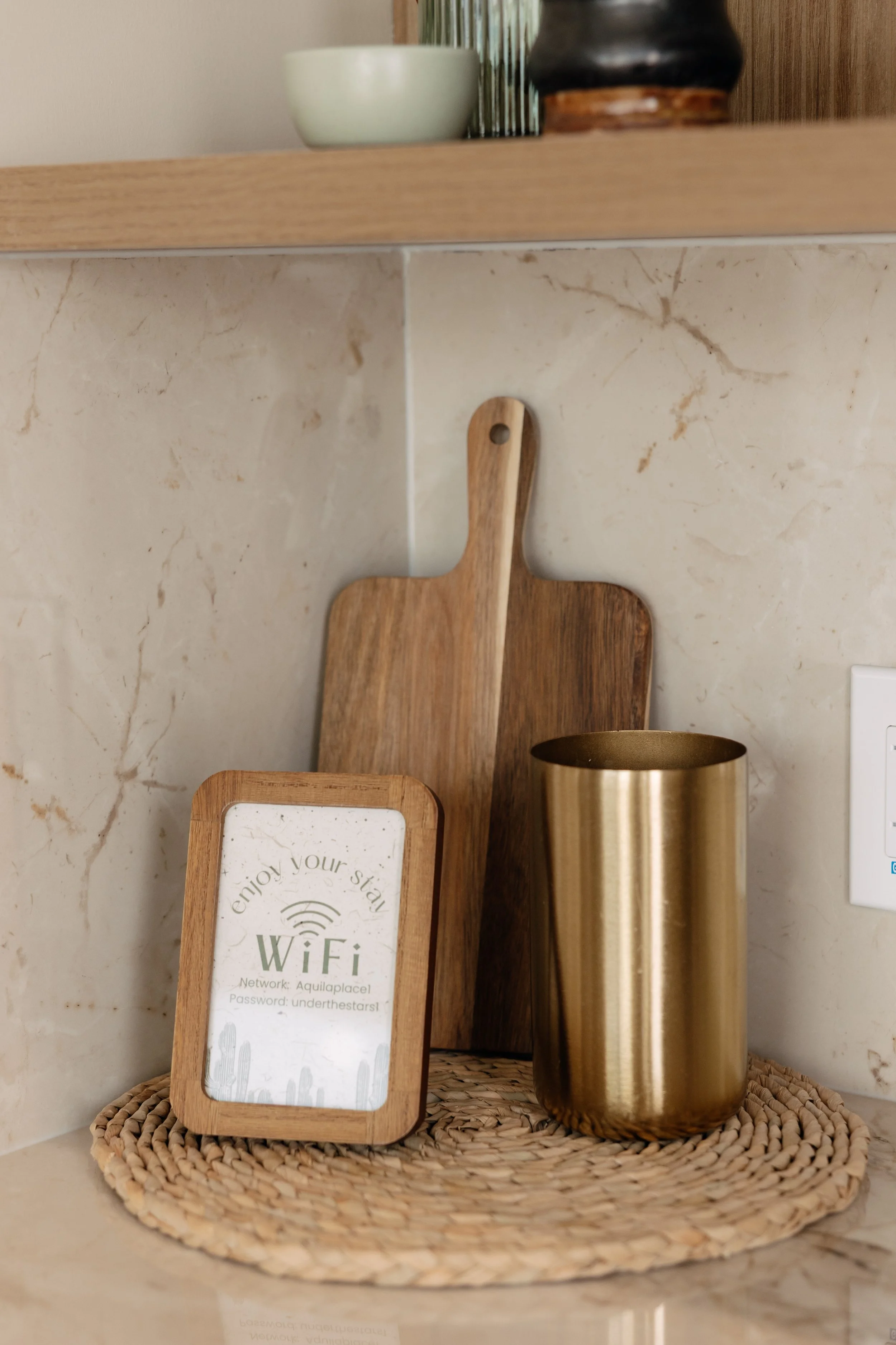 Decorative kitchen counter with a WiFi sign in a wooden frame, a gold metallic cup, a woven placemat, and two wooden cutting boards leaning against the wall.